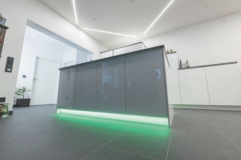 Nowoczesna kuchnia z szaro-błyszczącą wyspą, zielonym oświetleniem LED przy podłodze, otoczona białym, minimalistycznym wnętrzem.