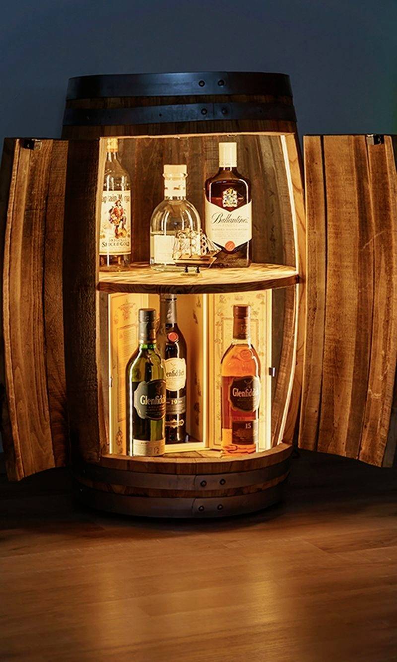 Drewniania beczka z otwartymi drzwiczkami ukazuje różne butelki alkoholu, w tym whisky i gin, ułożone na półce wewnątrz, oświetlone ciepłym światłem.