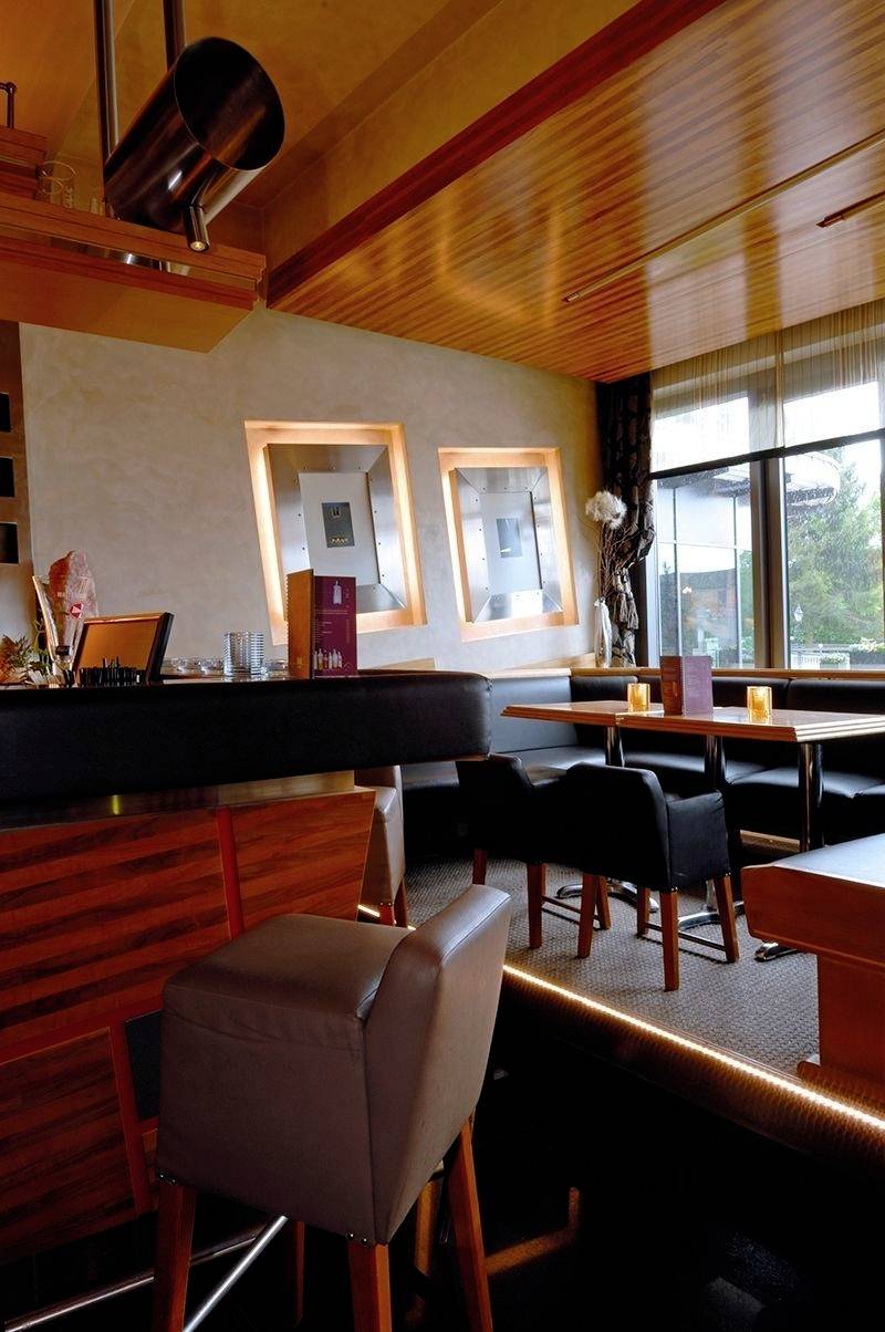 Przytulna kawiarnia z drewnianą dekoracją, nowoczesnymi meblami i dużymi oknami wpuszczającymi światło dzienne. Stoliki są przygotowane dla gości.