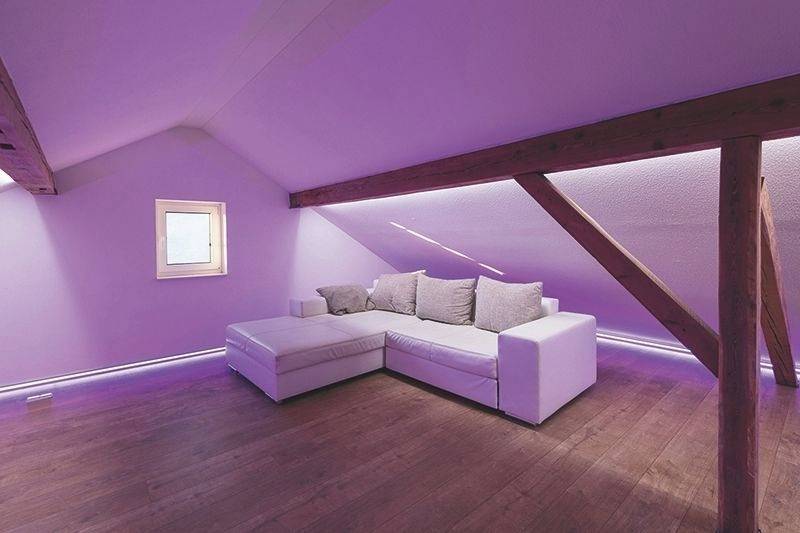 Minimalistyczny pokój na poddaszu z białą narożną sofą, drewnianą podłogą i fioletowym oświetleniem. Widoczne jest małe okno.
