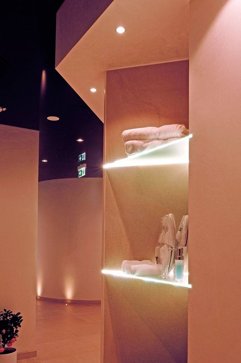 Oświetlone wnętrze spa z ręcznikami i produktami pielęgnacyjnymi na szklanych półkach, ciepłe światło ambientowe, widoczny znak wyjścia awaryjnego.