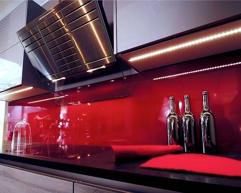 Nowoczesna kuchnia z błyszczącą czerwoną ścianą ochronną, stalowym wyciągiem kuchennym i dekoracyjnymi butelkami na czarnym blacie.