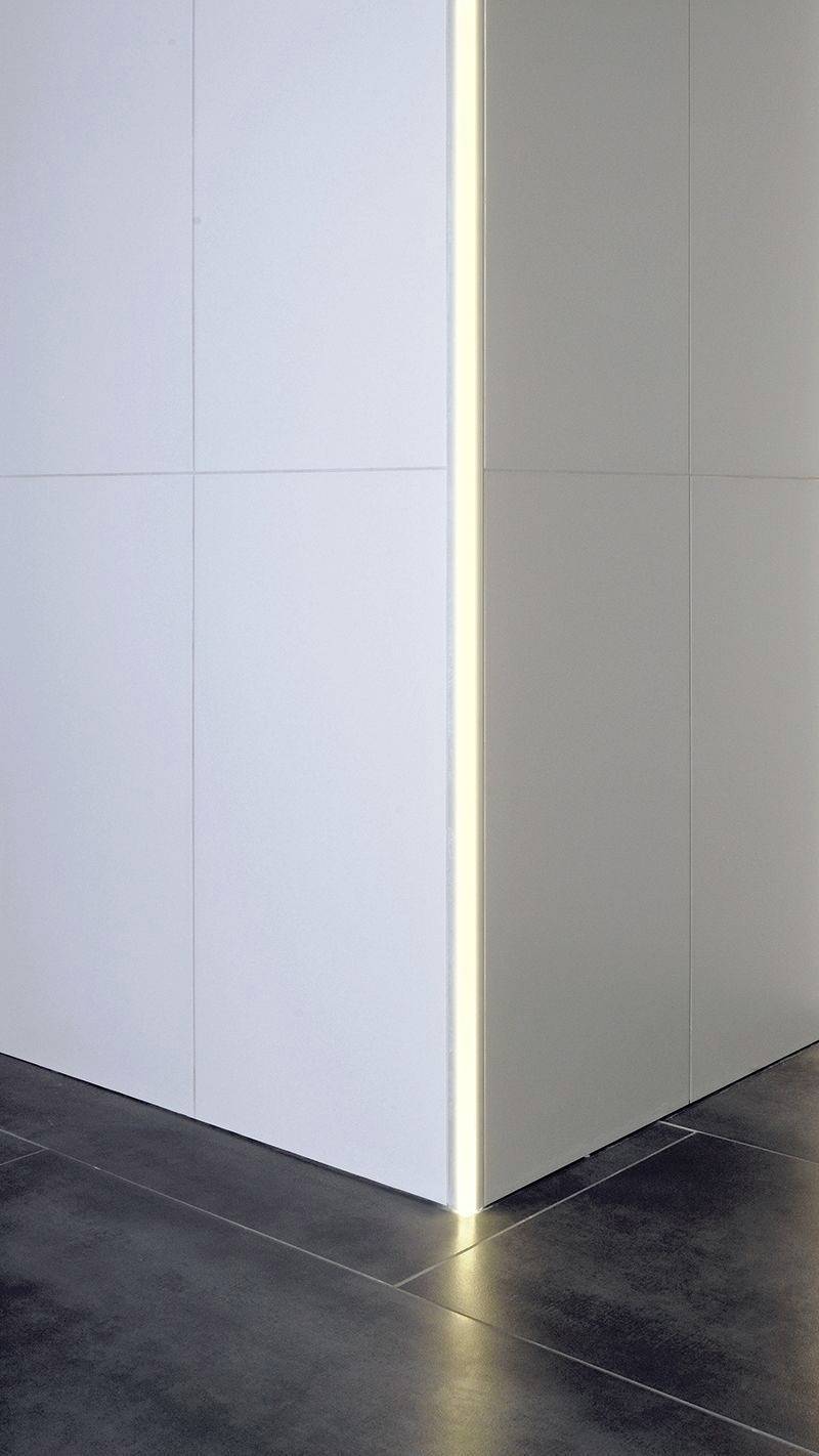Jasna ściana z subtelnym podświetleniem LED-owym wzdłuż krawędzi, na ciemnej podłodze z płytek. Minimalistyczny i nowoczesny design.