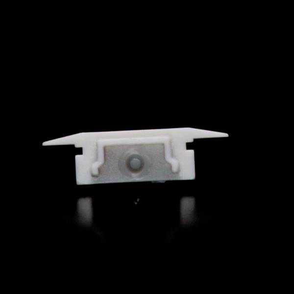 Mała, biała plastikowa część z dwoma bocznymi ramionami i centralnym okrągłym elementem, na czarnym tle.