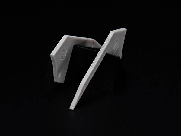 Dwie białe, plastikowe kliny na czarnym tle, które prawdopodobnie mogą być używane jako dystanse lub do mocowania.