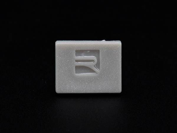 Prostokawisty, biały przycisk z wygrawerowanym, stylizowanym logo 'R' na czarnym tle.