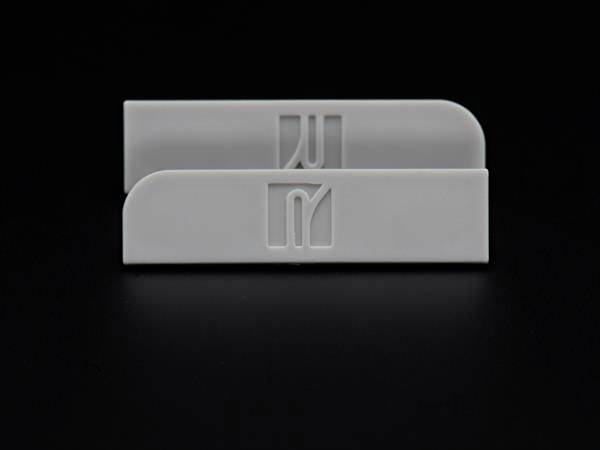 Dwa jasnoszare, prostokątne plastikowe elementy z zaokrąglonymi narożnikami na czarnym tle, oba z wygrawerowanym stylizowanym logo lub symbolem.