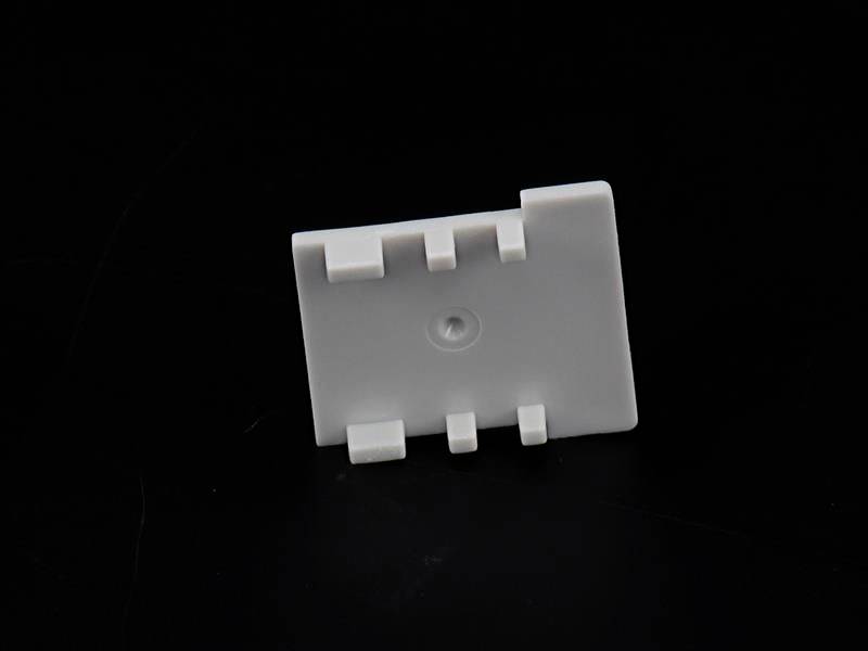 Biała plastikowa część z wieloma wycięciami i otworem pośrodku, na czarnym tle.