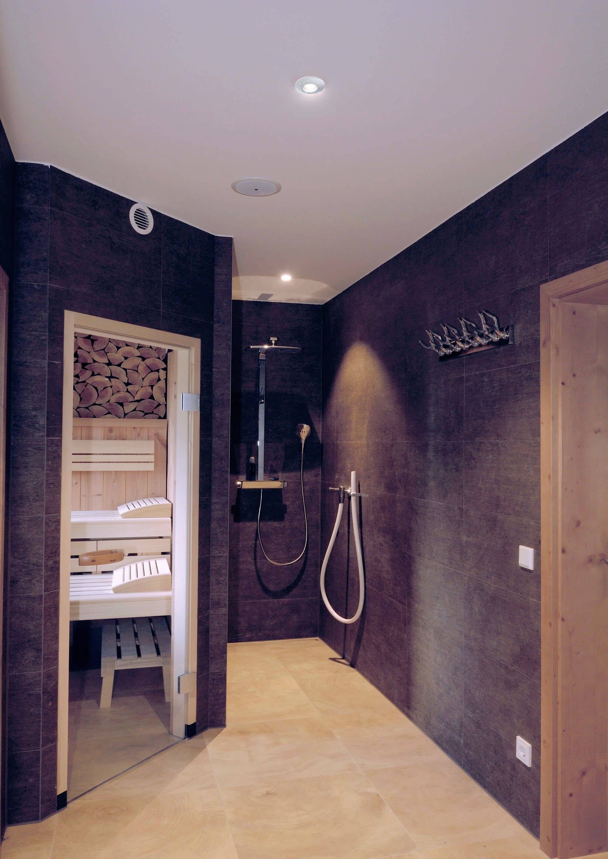 'Prysznic i sauna w nowoczesnej strefie kąpielowej z ciemnymi ścianami i jasną podłogą. Po lewej stronie widoczny jest stos drewna opałowego.'