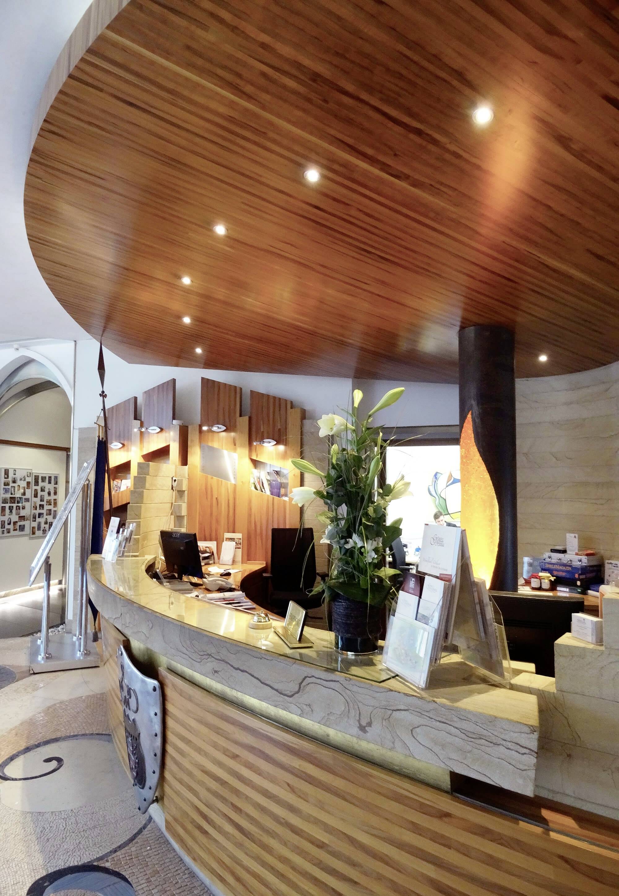 Recepcja hotelowa z drewnianym sufitem. Nowoczesne oświetlenie, dekoracje roślinne i komputer na ladzie.