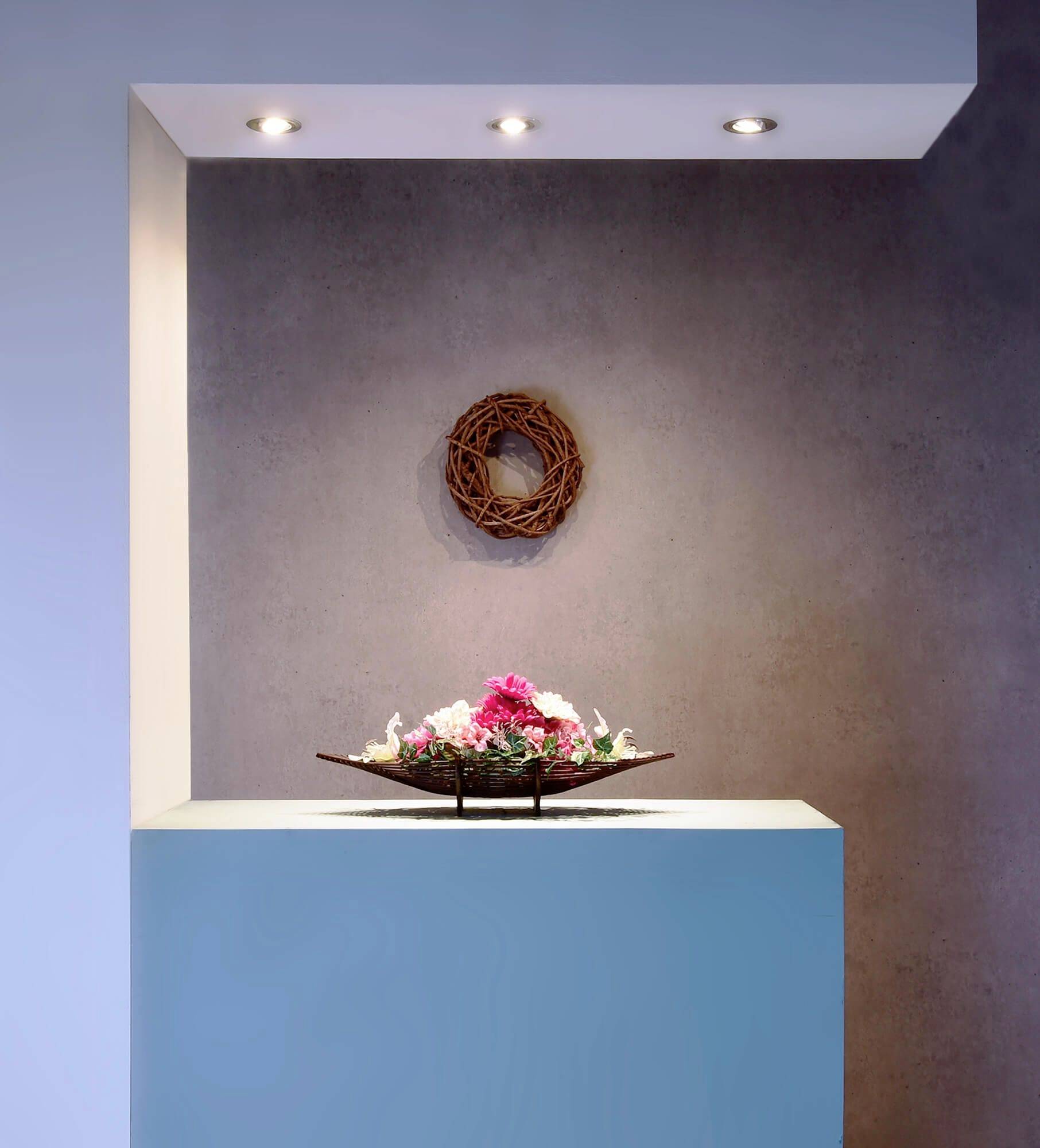 Dekoracyjna kompozycja kwiatowa w misce stoi na turkusowym podeście przed szarą ścianą, na której wisi pleciony wianek.