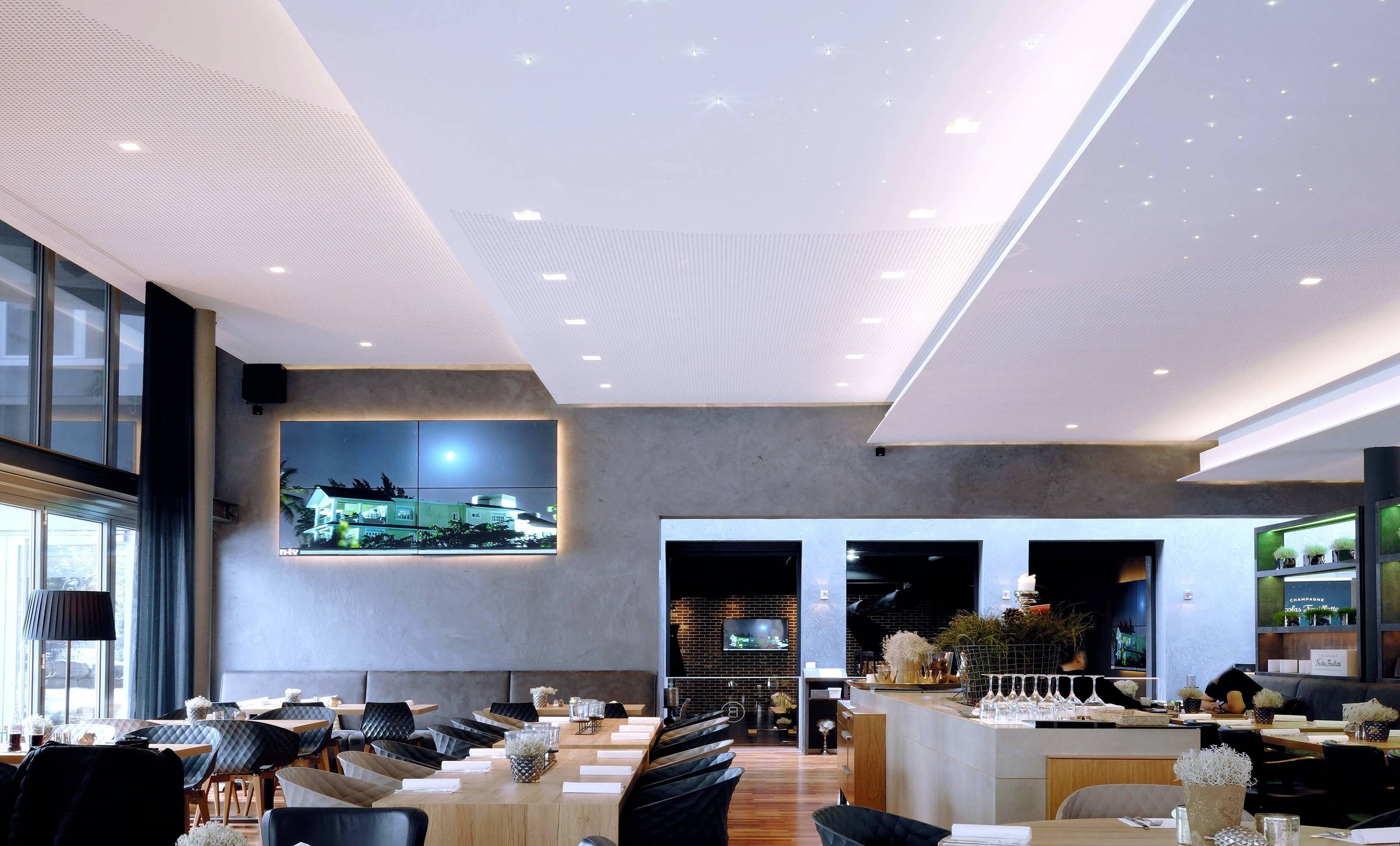 Elegancka restauracja z nowoczesnymi meblami, drewnianymi detalami i podświetlanym sufitem. Na ścianie wisi obraz przedstawiający nocny widok.