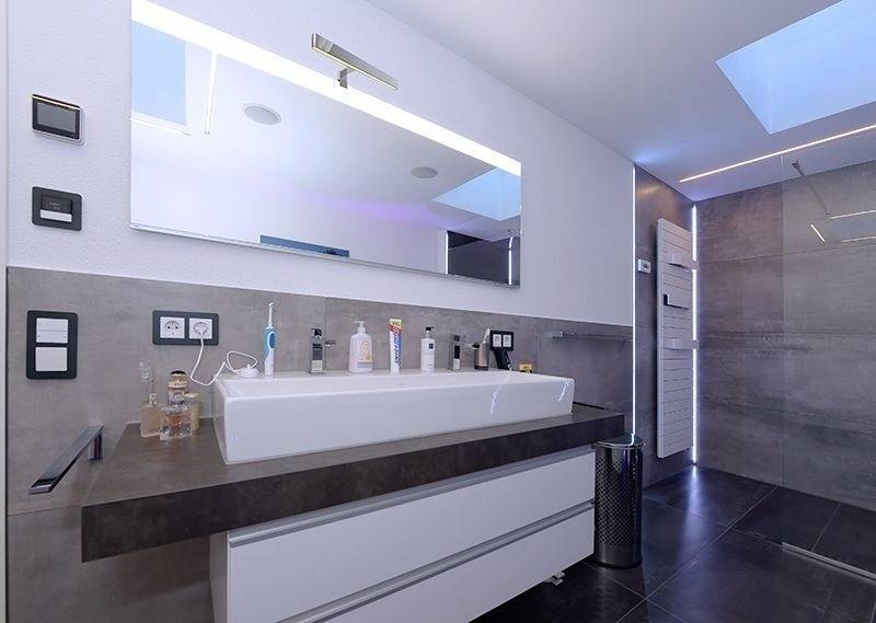 Nowoczesna łazienka z dużym lustrem, umywalką, kilkoma produktami pielęgnacyjnymi i strefą prysznica. Jasne oświetlenie przez okno w suficie.