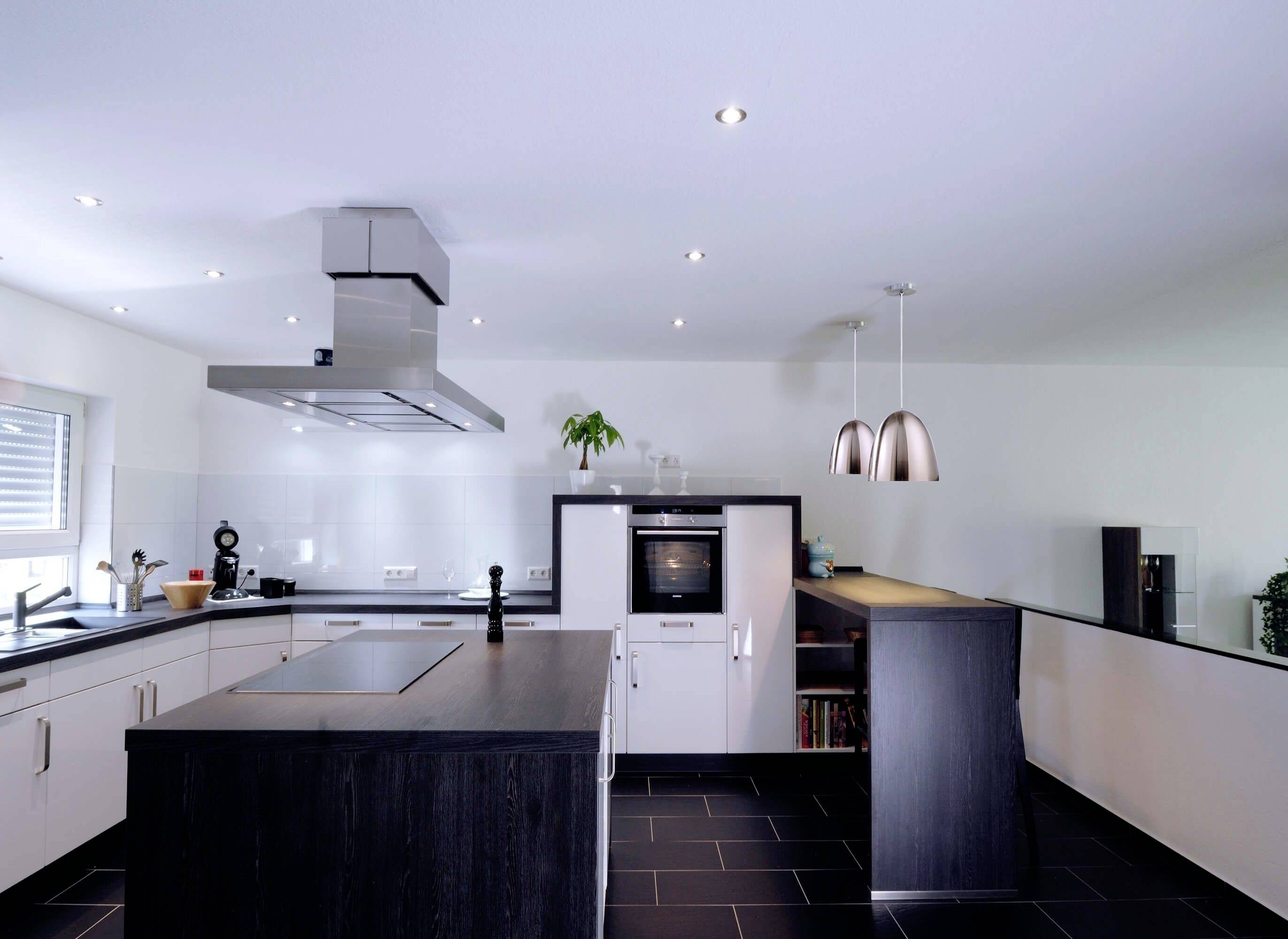 Nowoczesna kuchnia z centralną wyspą kuchenną, czarnym blatem, białymi szafkami i stalową wyciągową okapem. Wiszące lampy i piekarnik przy tylnej ścianie.