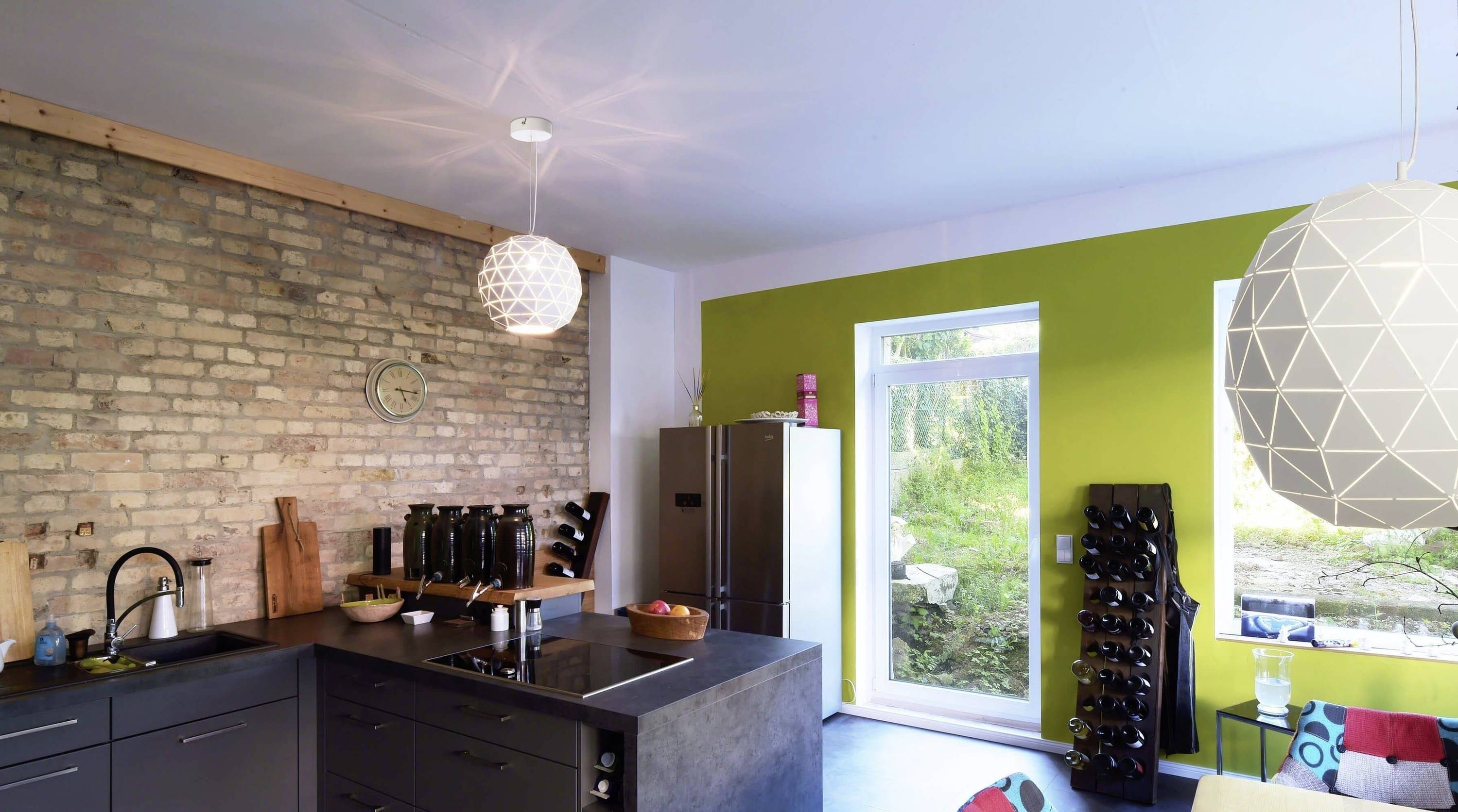 Nowoczesna kuchnia z ścianą z czerwonej cegły, wolnostojącą lodówką, wyspą kuchenną i zieloną ścianą akcentową. Duże okna oferują widok na ogród.
