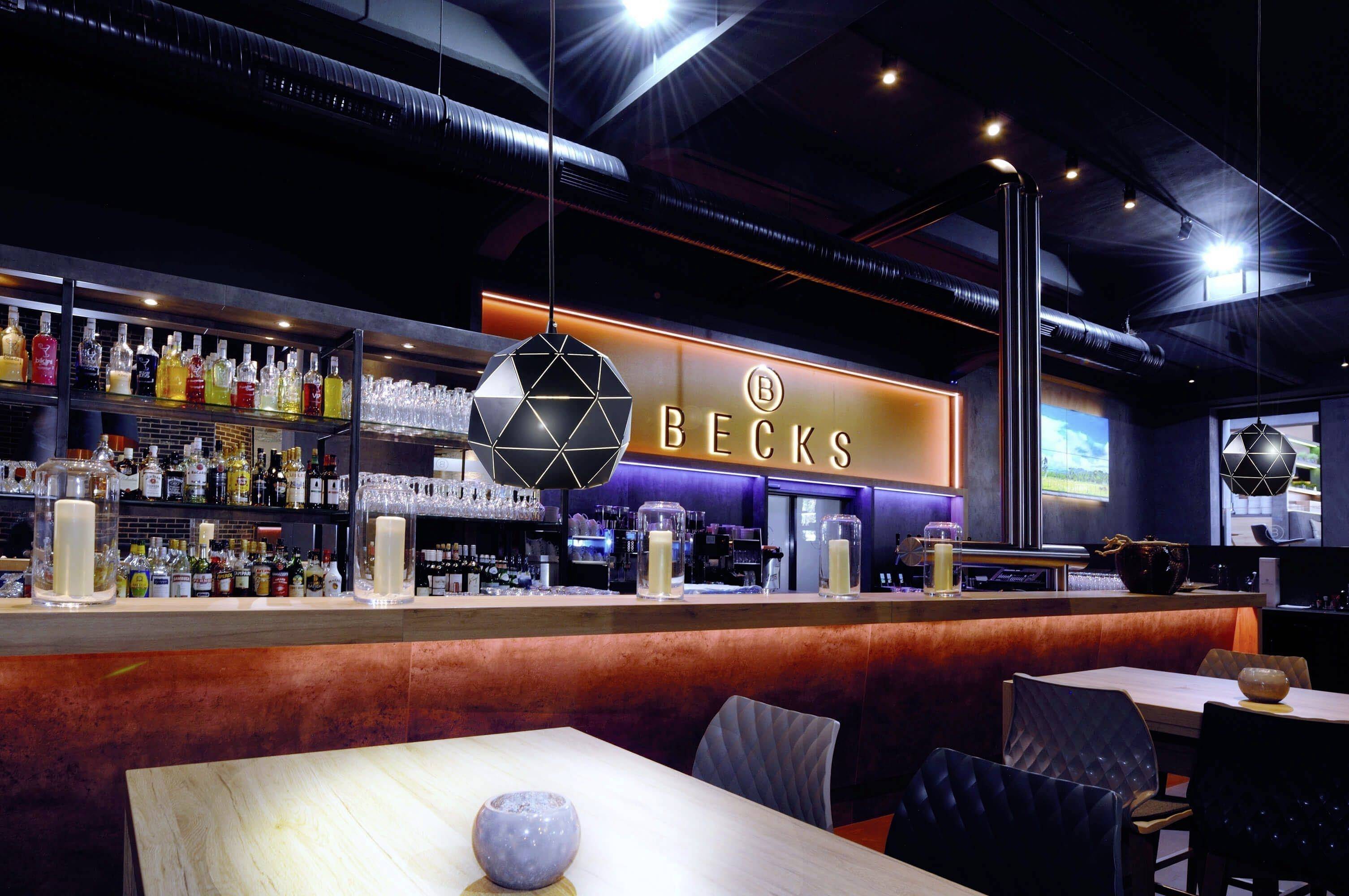Nowoczesny bar z podświetlanym barem i półkami pełnymi butelek w tle. Neonowy napis 'Becks' na ścianie.