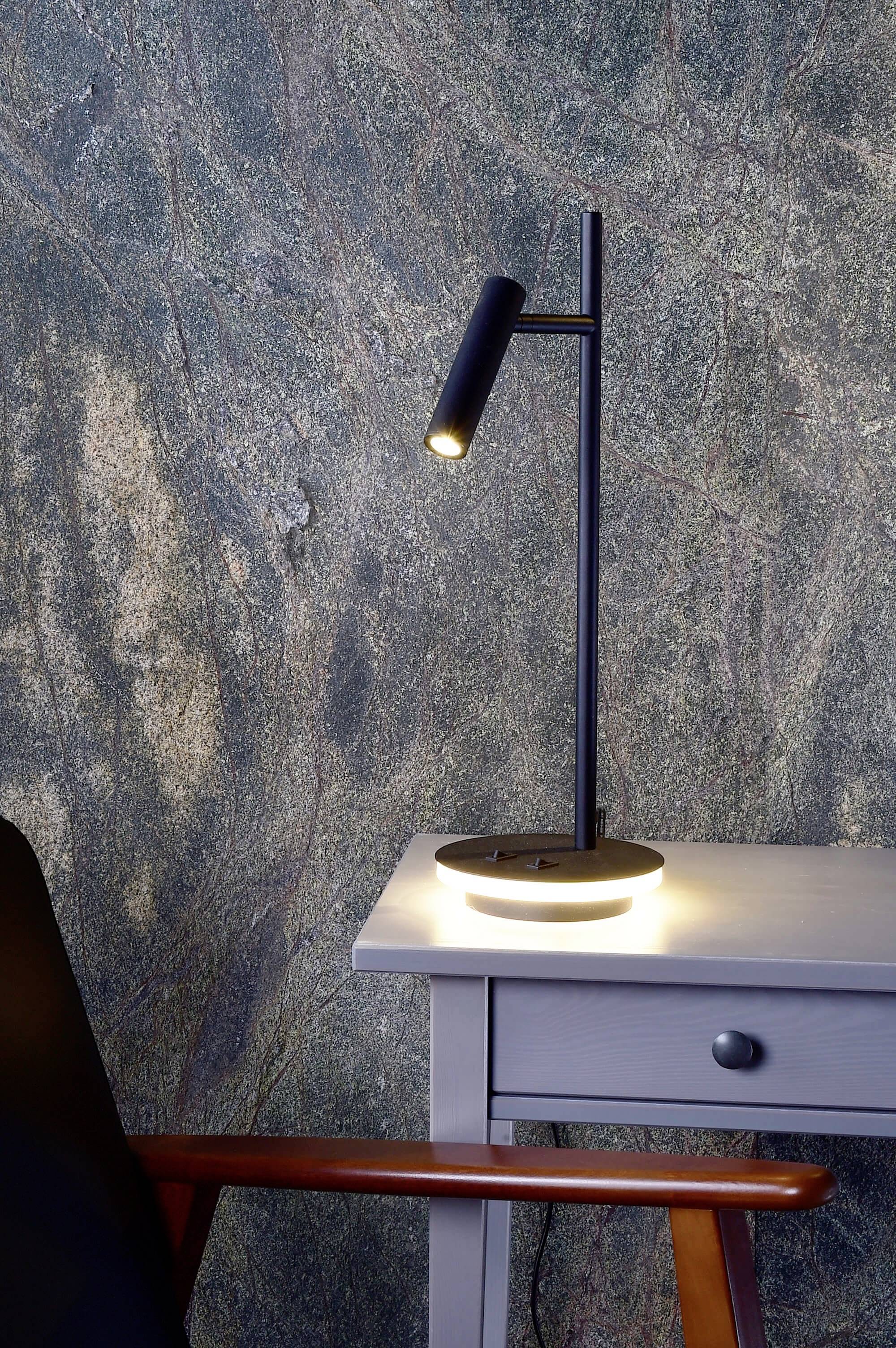 Smukła, czarna lampa stołowa świeci na szarym drewnianym stole przed ścianą o fakturowej powierzchni. Obok stoi krzesło.