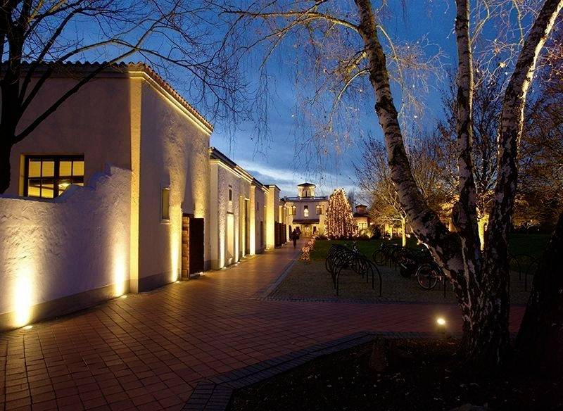 Oświetlona droga biegnie wzdłuż białych budynków otoczonych drzewami. Panuje wieczorowy nastrój, a niebo jest bezchmurne.