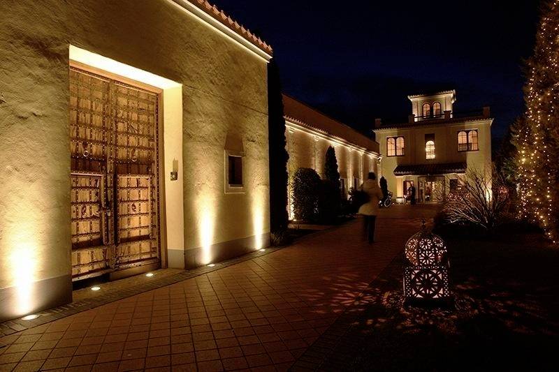 Historyczny budynek oświetlony nocą, z wielkimi drewnianymi drzwiami i latarnią na pierwszym planie, ludzie spacerujący w tle wzdłuż drogi.