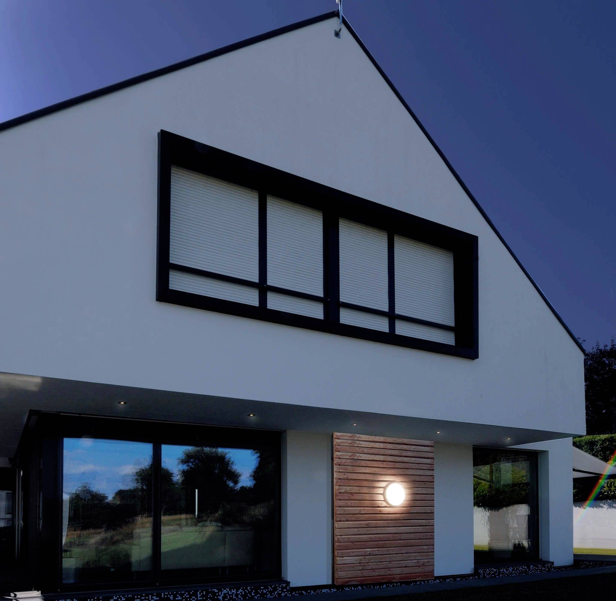 Nowoczesny dom jednorodzinny z białą elewacją, dużymi oknami i wejściem wyłożonym drewnem. Na tle czystego, błękitnego nieba.