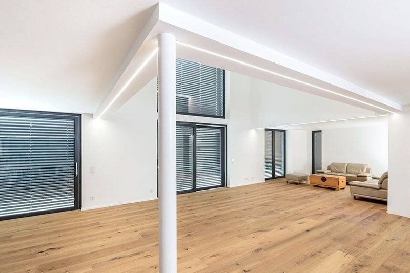 Nowoczesny salon z drewnianą podłogą, białymi ścianami i dużymi oknami z żaluzjami. Beżowa kanapa stoi w prawym rogu.