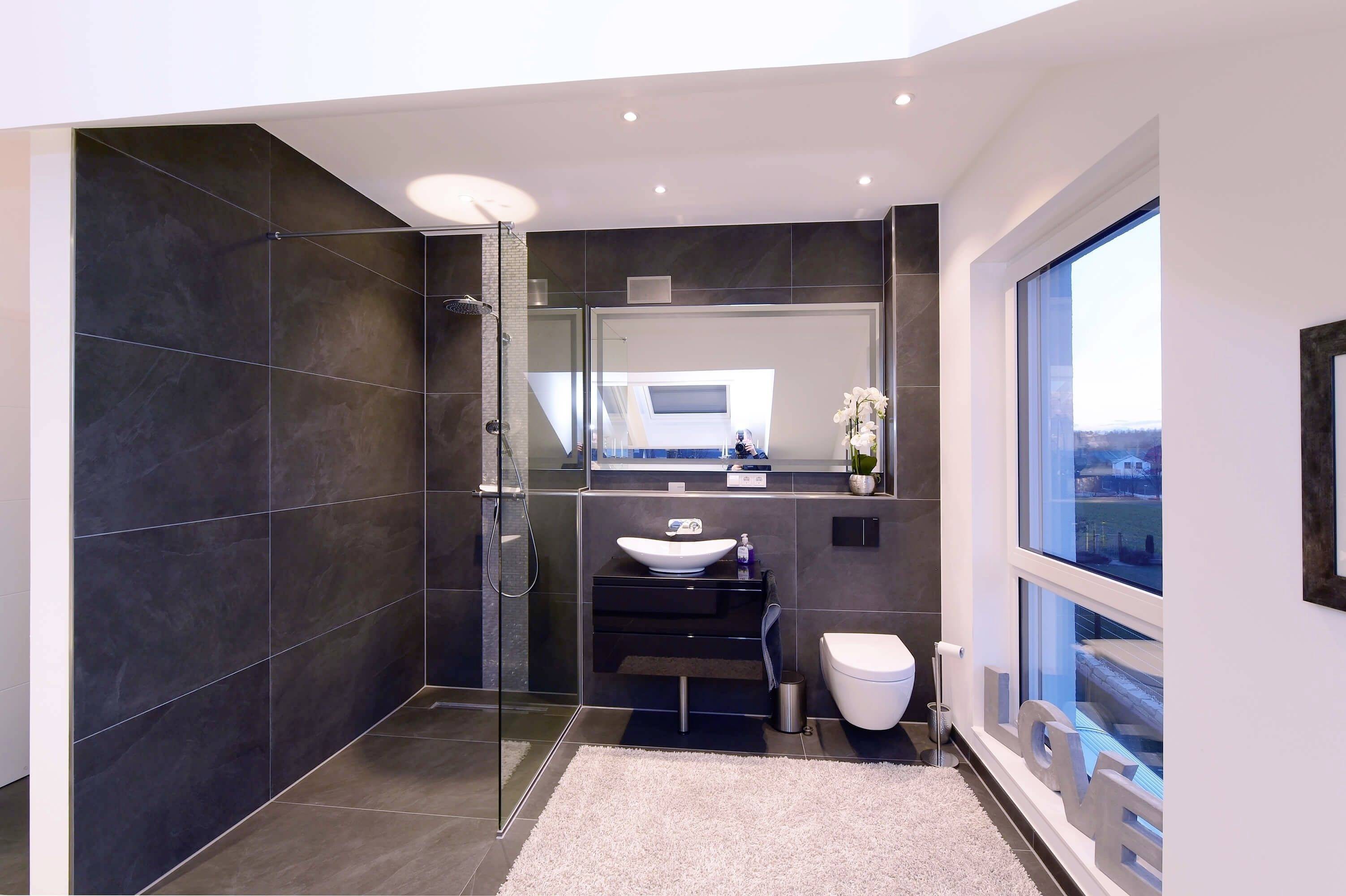 Nowoczesna łazienka ze szklaną kabiną prysznicową, umywalką, toaletą i dużym oknem. Jasne ściany kontrastują w stylowy sposób z ciemnymi płytkami.