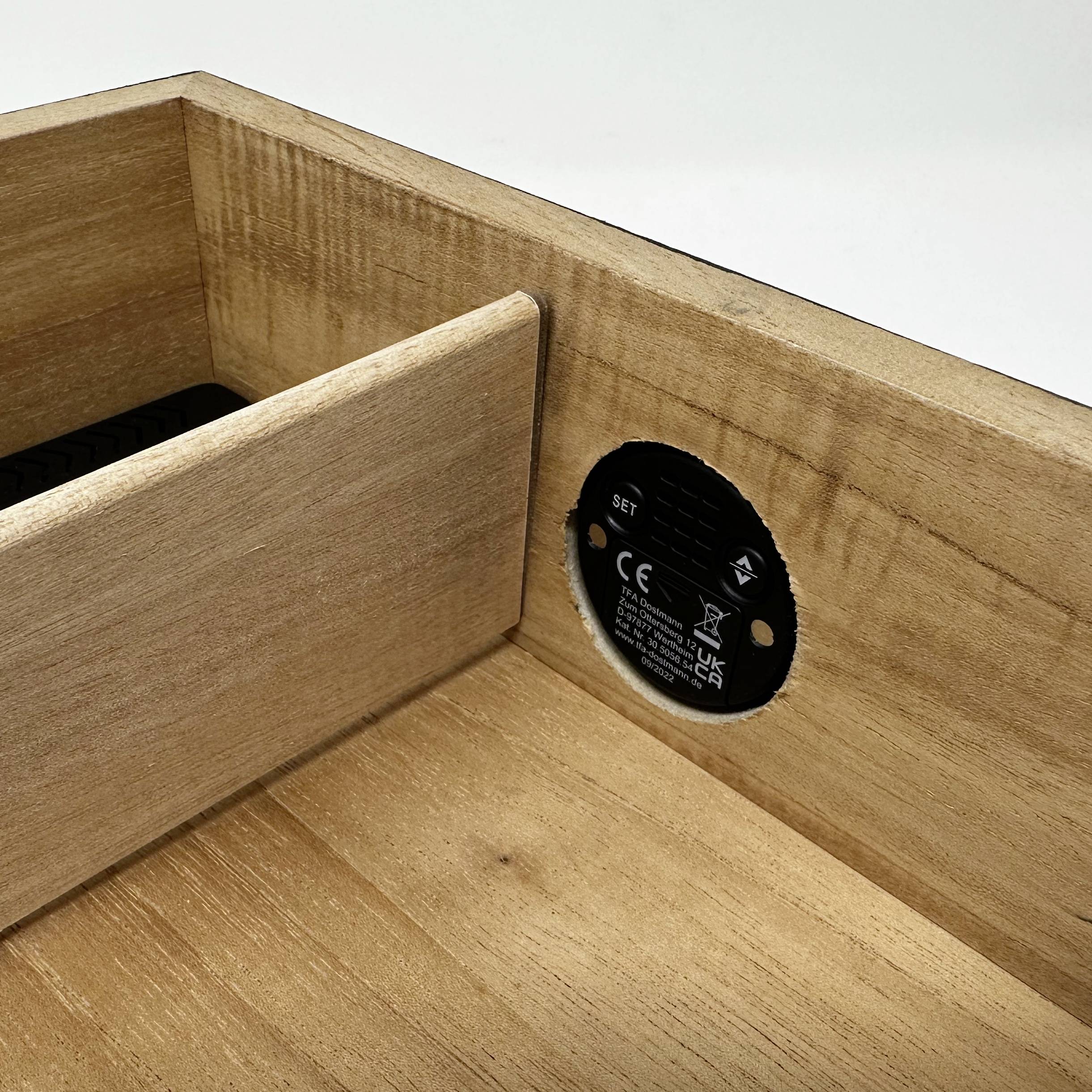 Drewniana szuflada z okrągłą, czarną pokrywą baterii po wewnętrznej stronie. Na tylnej ścianie szuflady widoczna wąska szczelina.