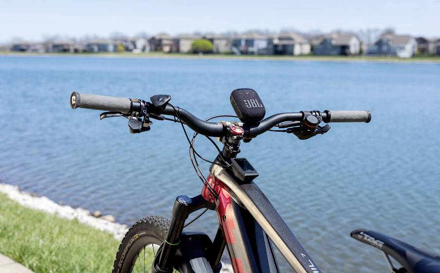 Rower z głośnikiem JBL zamocowanym na kierownicy stoi nad jeziorem, otoczony trawą i rzędem domów w tle.