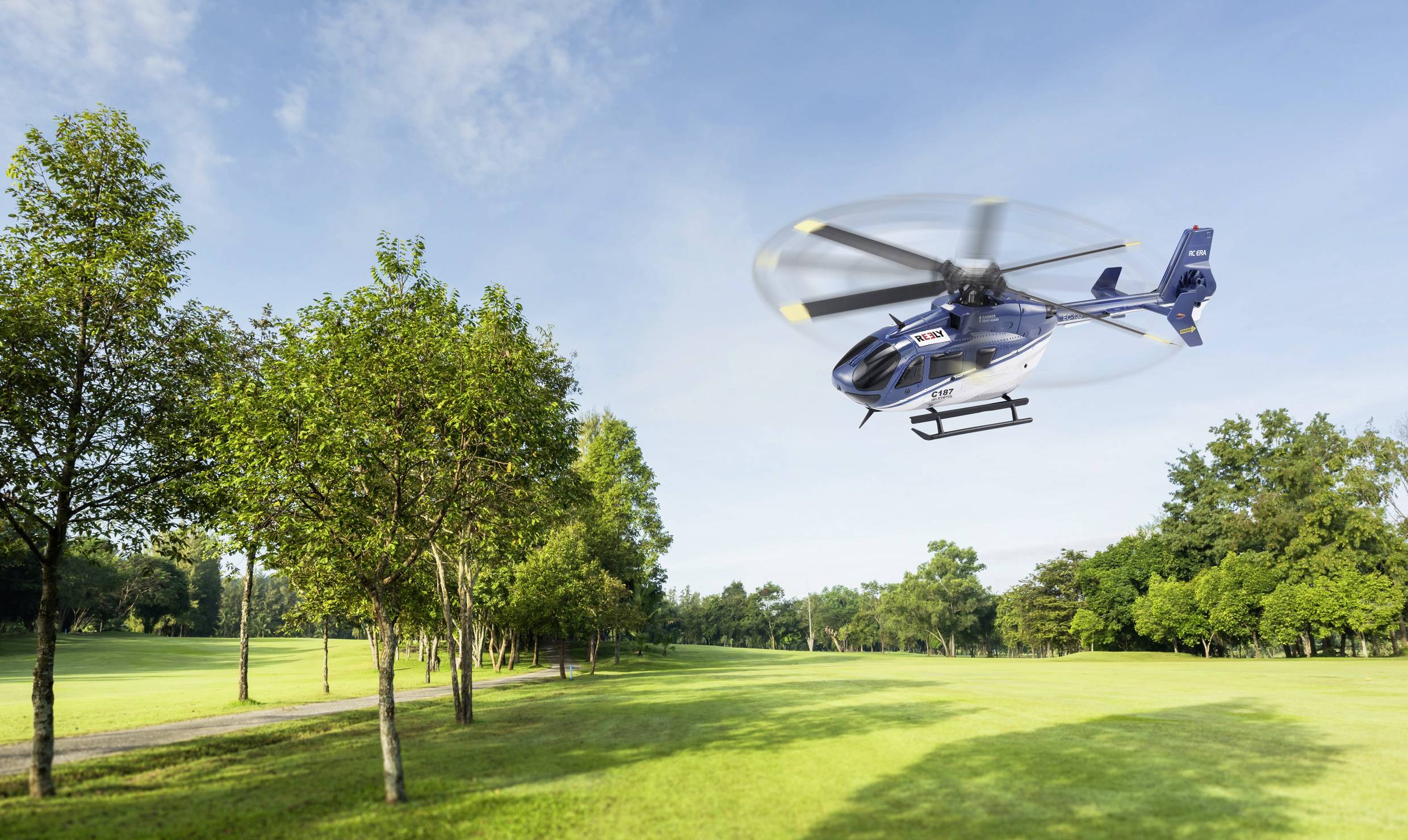 Niebieski helikopter leci nad zieloną łąką z drzewami. W tle widać bezchmurne niebo.