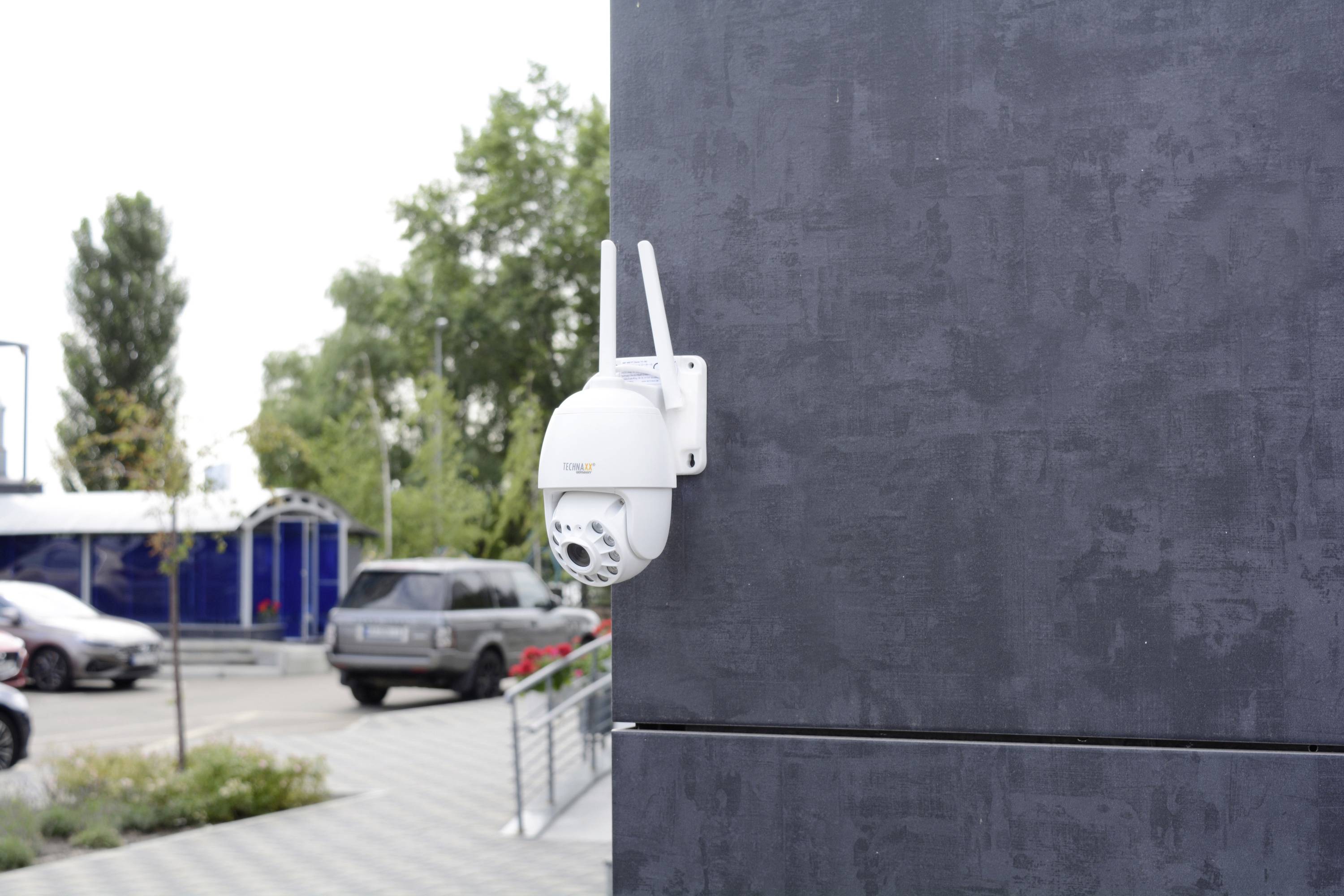 Kamera monitoringu zamocowana na szarej ścianie, skierowana na parking z samochodami i drzewami w tle.