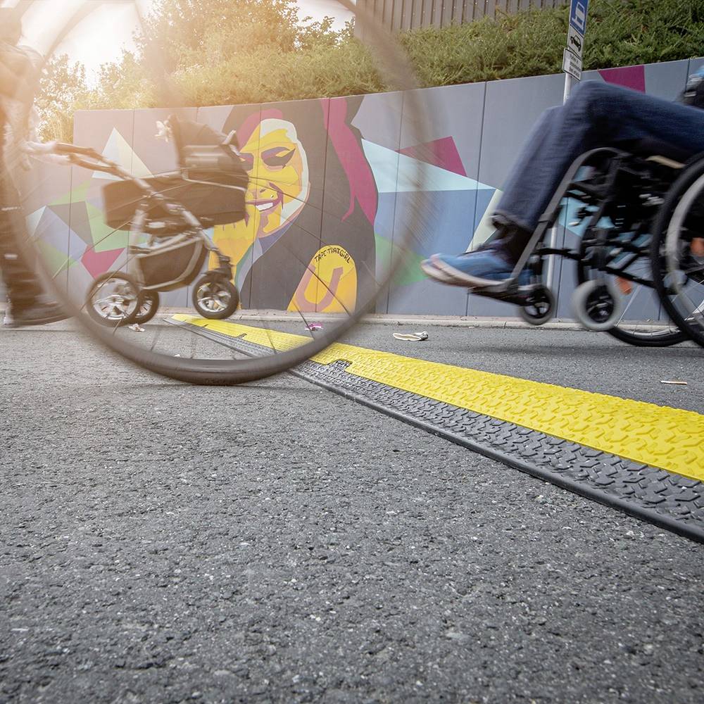 Ulica z drogą rowerową, żółtymi oznaczeniami oraz ludźmi z wózkiem dziecięcym i wózkiem inwalidzkim, na tle kolorowej malowanej ściany w tle.