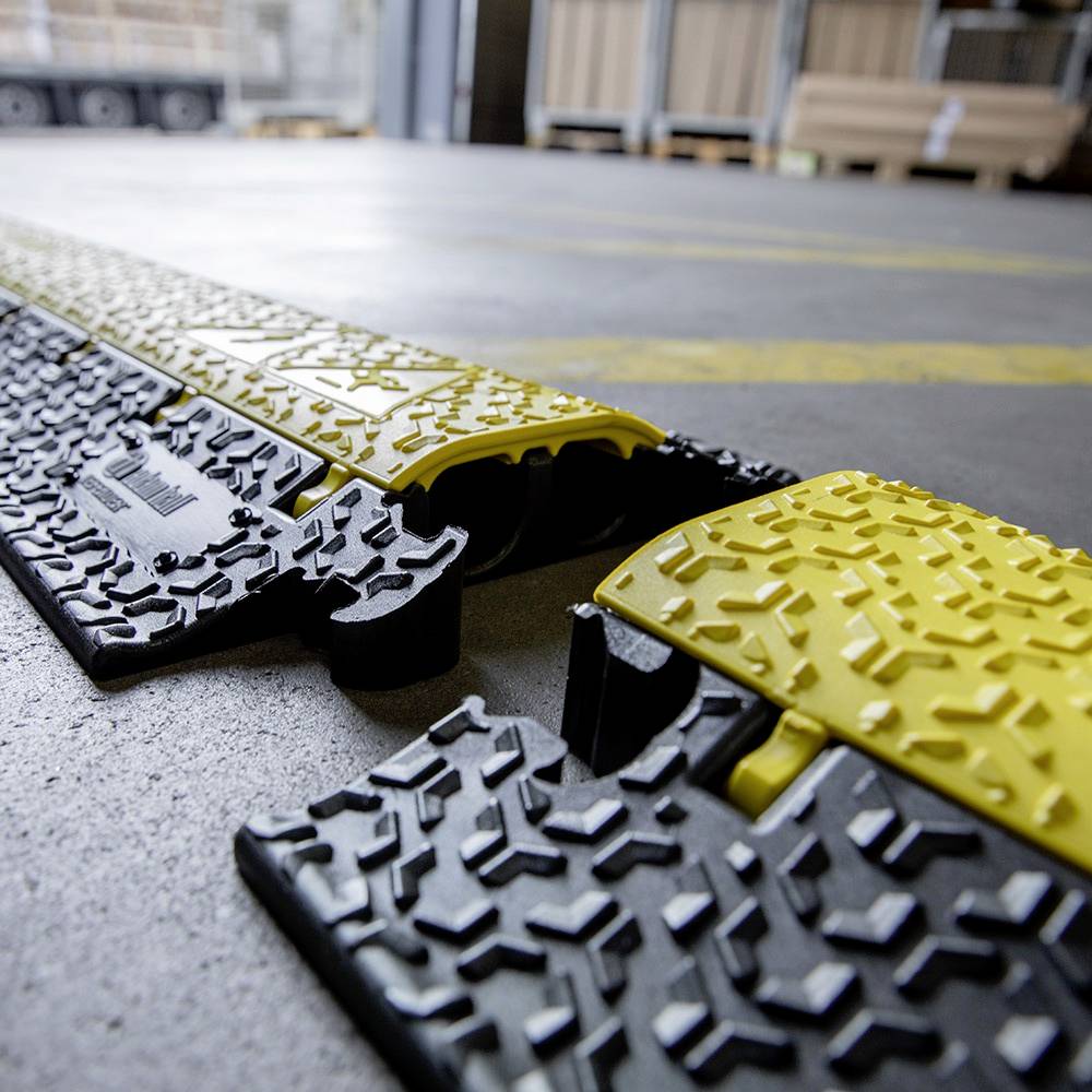 Żółto-czarny kanał kablowy biegnie przez podłogę magazynu. Kanał zabezpiecza przewody i eliminuje ryzyko potknięcia się w środowisku przemysłowym.