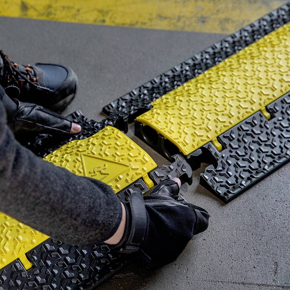 Osoba montuje żółte i czarne osłony kabli na podłodze. Płyty posiadają symbol ostrzegawczy.