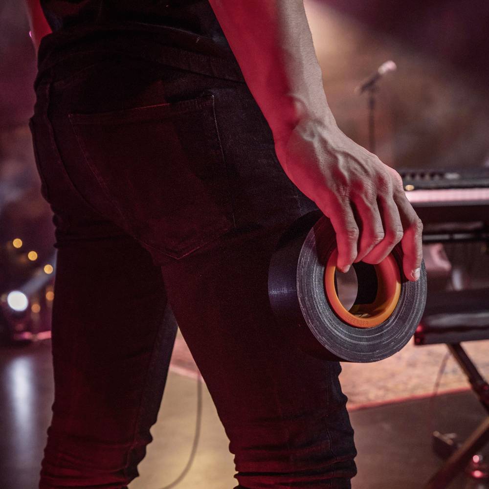 Osoba trzyma taśmę klejącą w ręku, stoi na scenie; w tle niewyraźnie widać mikrofon i sprzęt muzyczny.