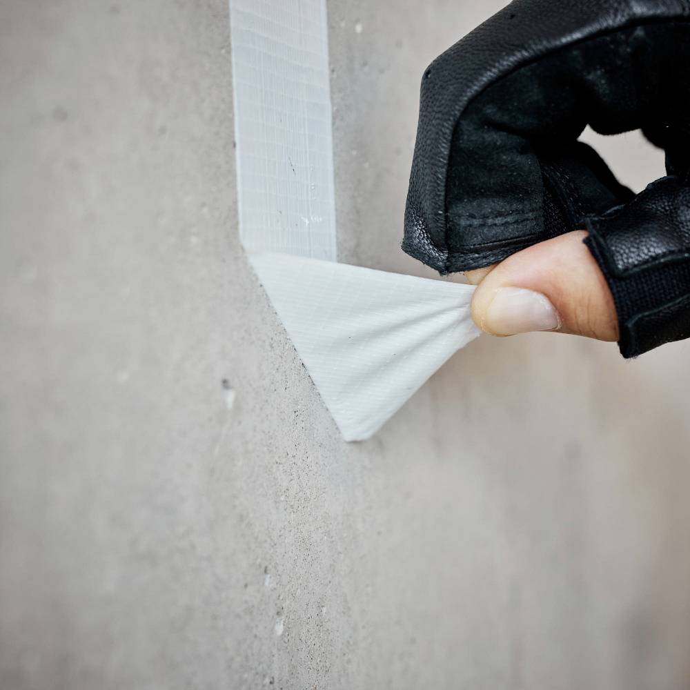 Dłoń w rękawicy odklejająca taśmę klejącą od betonowej ściany.