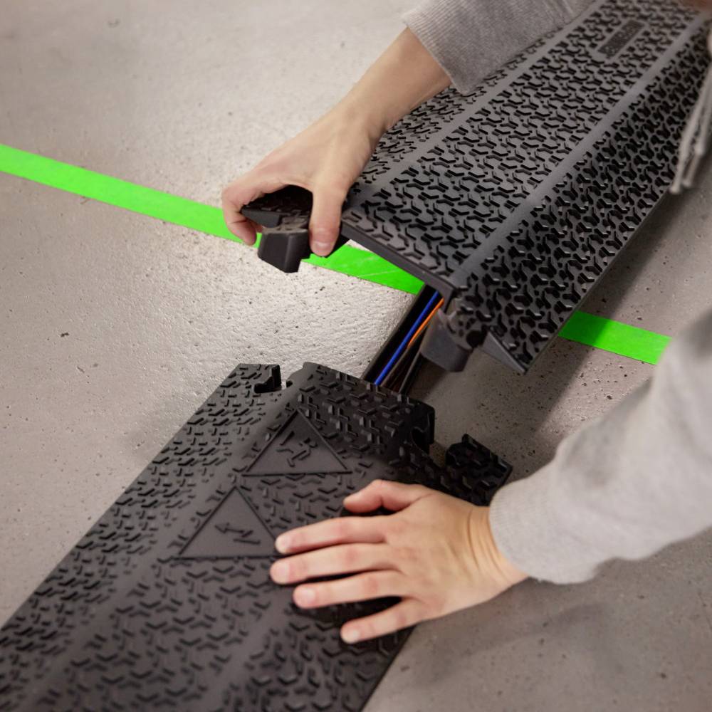 Osoba instaluje osłonę kabli z przewodami gumowymi na podłodze, zielona linia wskazuje granicę. Ręce precyzyjnie umieszczają osłonę.