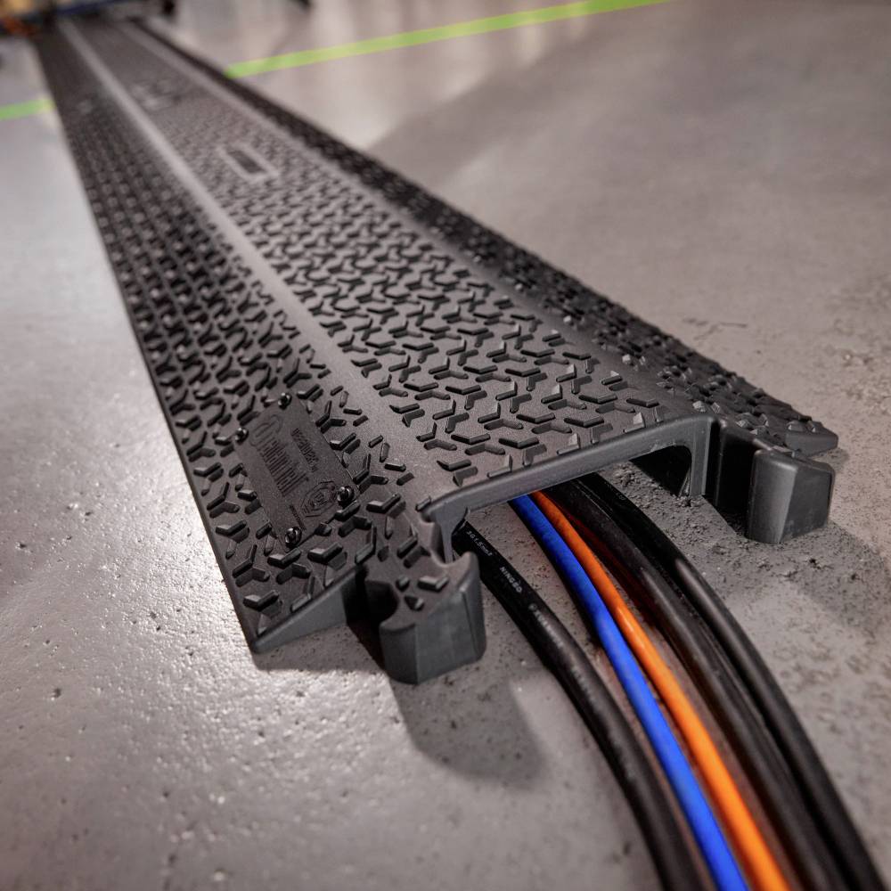 Naziemna osłona kabli: Czarna rampa z wieloma kablami pod spodem, zapewniająca ochronę i porządek w środowisku przemysłowym.