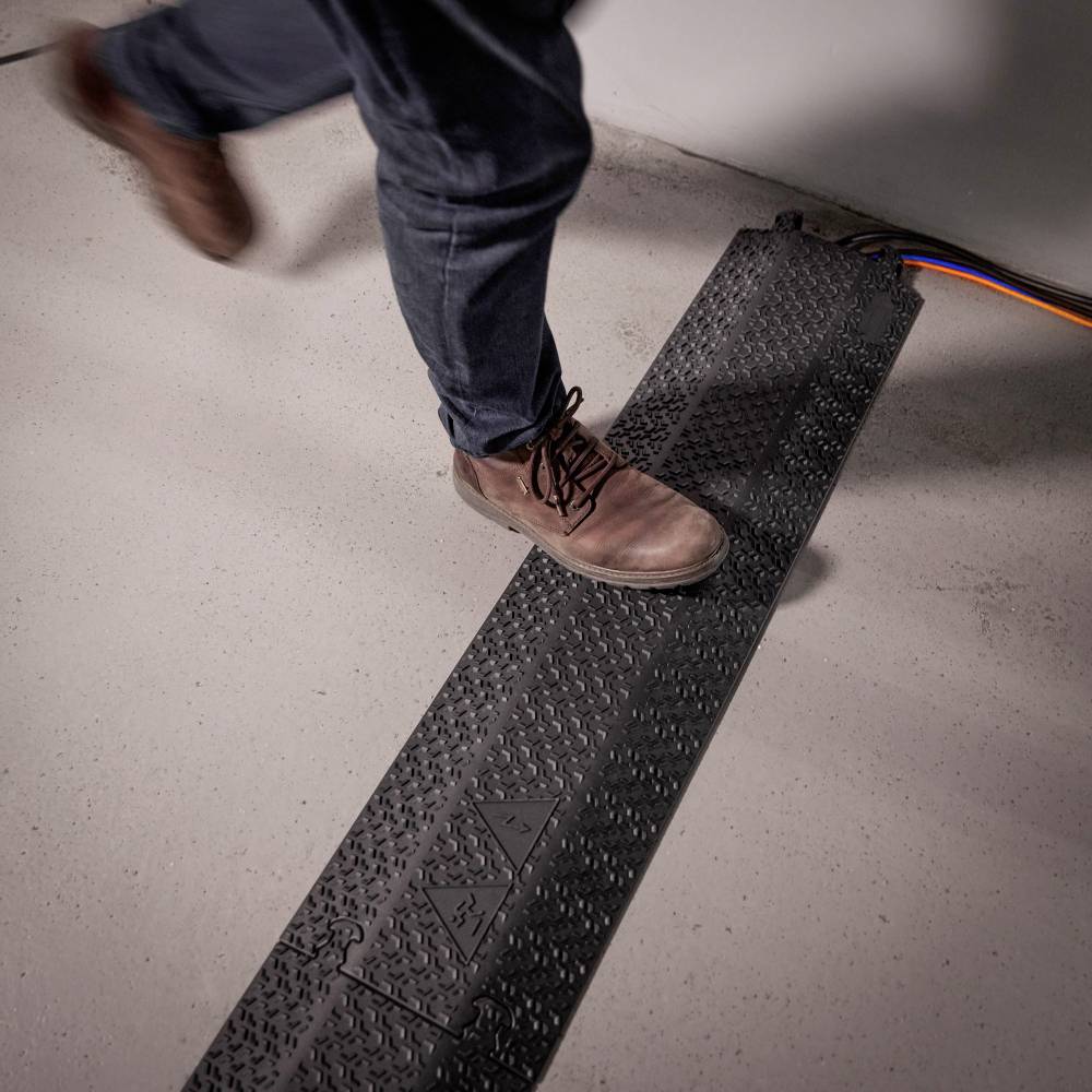 Osoba przechodzi przez czarną osłonę kabla na szarym podłożu, aby uniknąć niebezpieczeństwa potknięcia. W tle widoczny jest kabel.