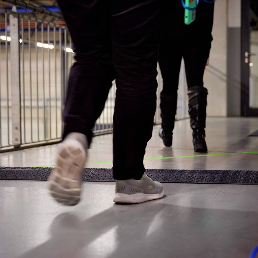 Osoba przechodzi przez korytarz wyłożony dywanem. W tle widoczne są metalowe poręcze.