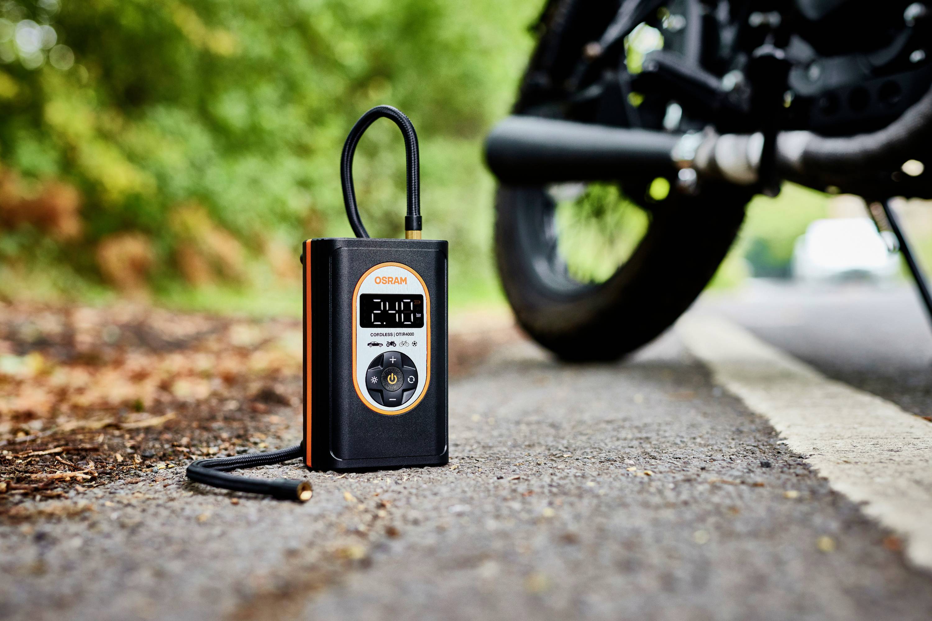 Przenośna sprężarka leży na drodze obok roweru. Wyświetlacz pokazuje '2.40'. W tle widoczne są drzewa i liście.