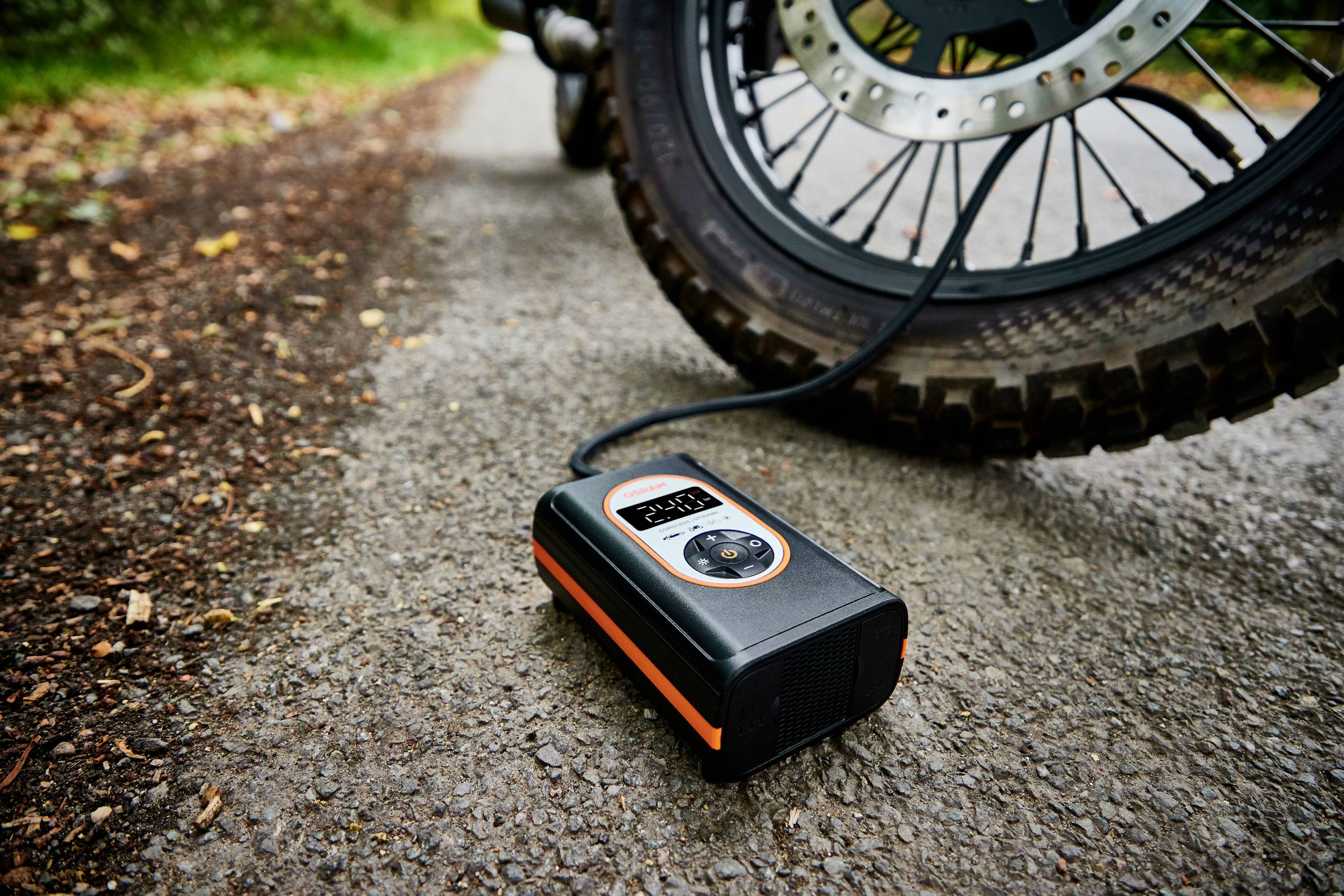 Przenośny kompresor do opon napełnia oponę motocykla. Na wyświetlaczu kompresora widać '2,2', a sam kompresor leży na asfaltowej drodze.