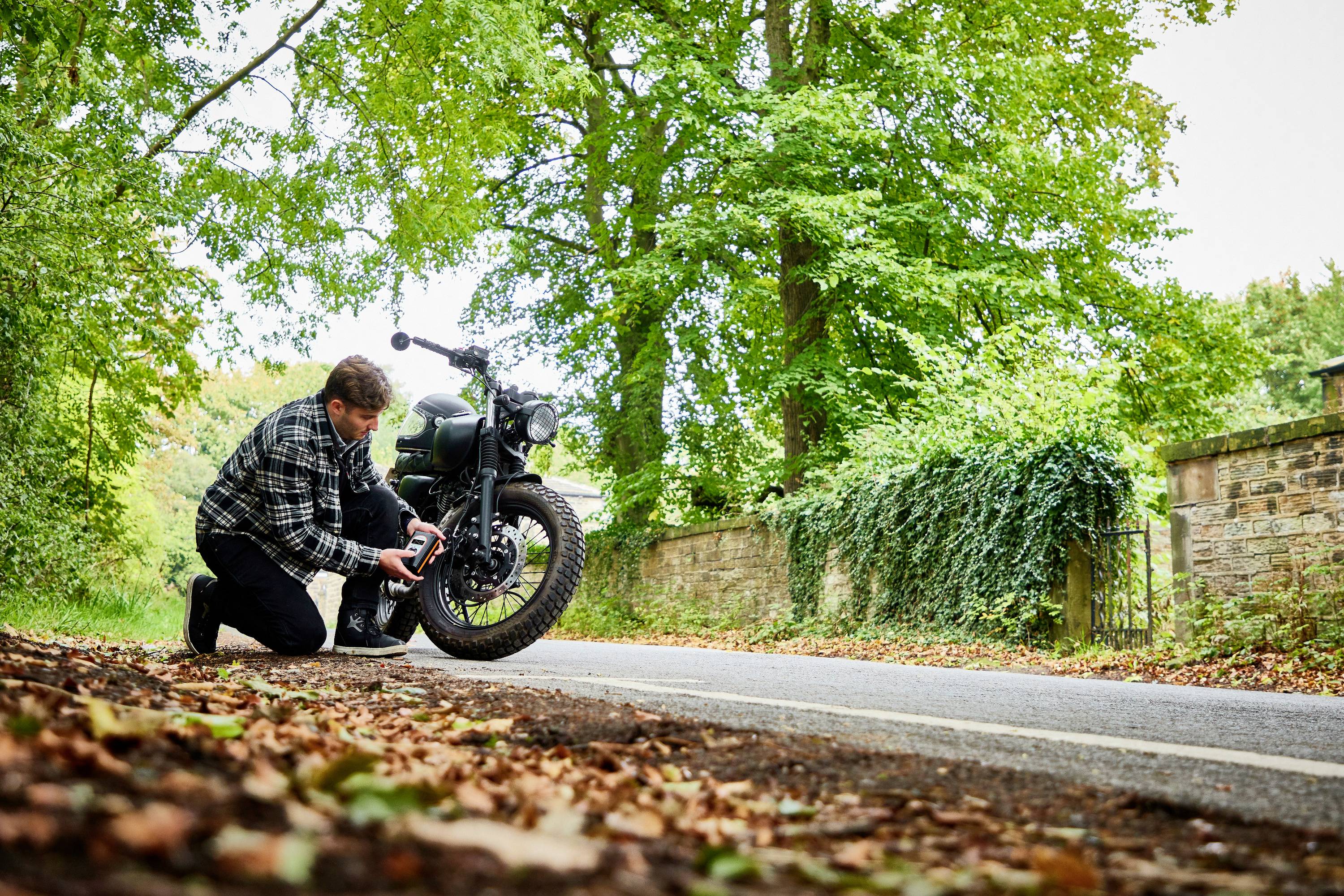 Mężczyzna klęczy obok motocykla przy drodze i sprawdza ciśnienie w oponach. W tle widoczne są drzewa i kamienny mur.
