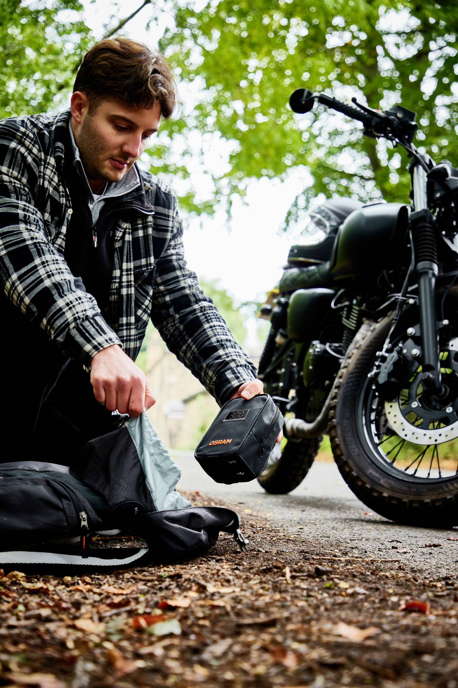 Mężczyzna klęczy obok otwartej torby na leśnej ścieżce i trzyma małe pudełko. W tle stoi motocykl.