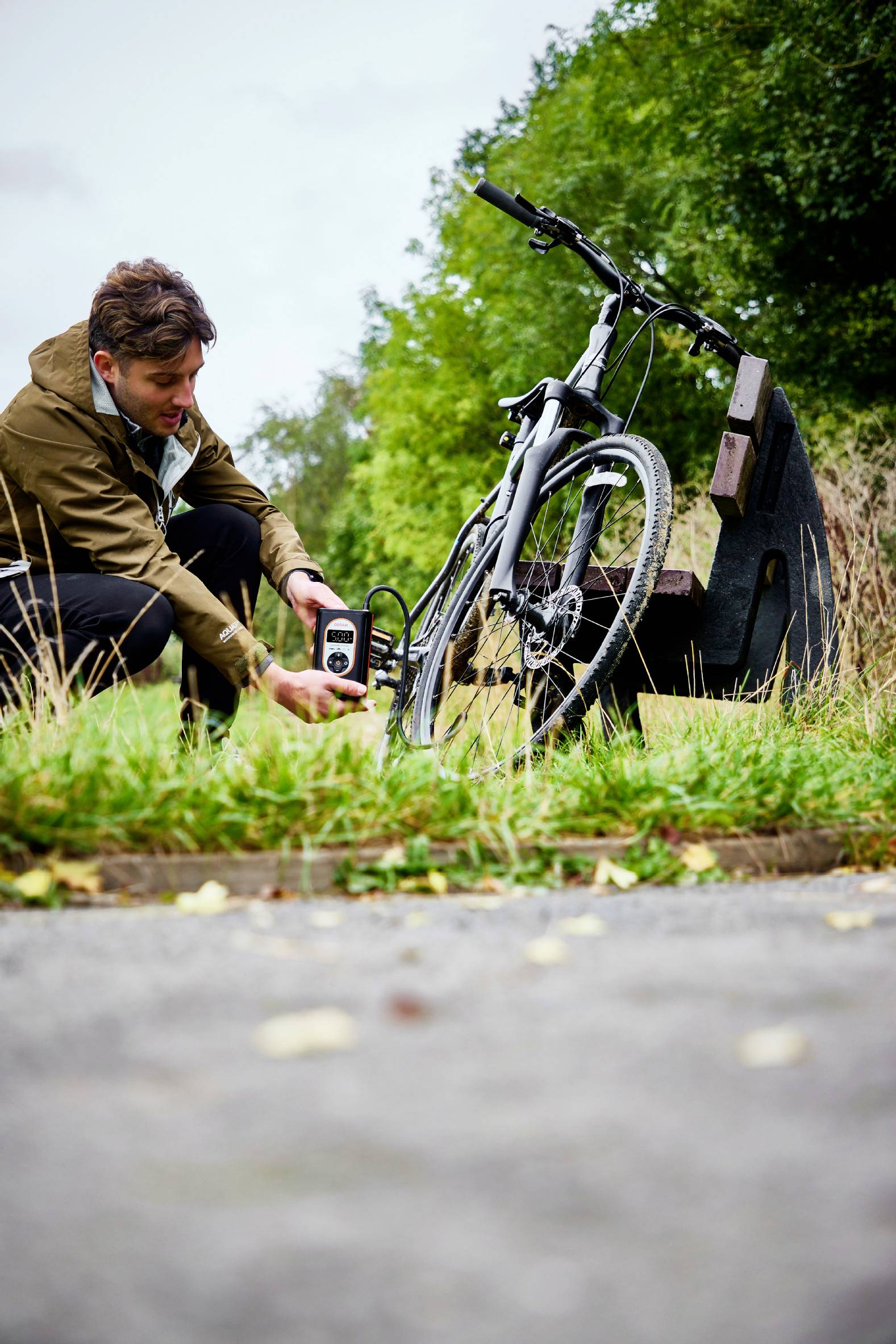 Osoba pompuje rower ręczną pompką na ścieżce. W tle widoczne są drzewa i ławka.