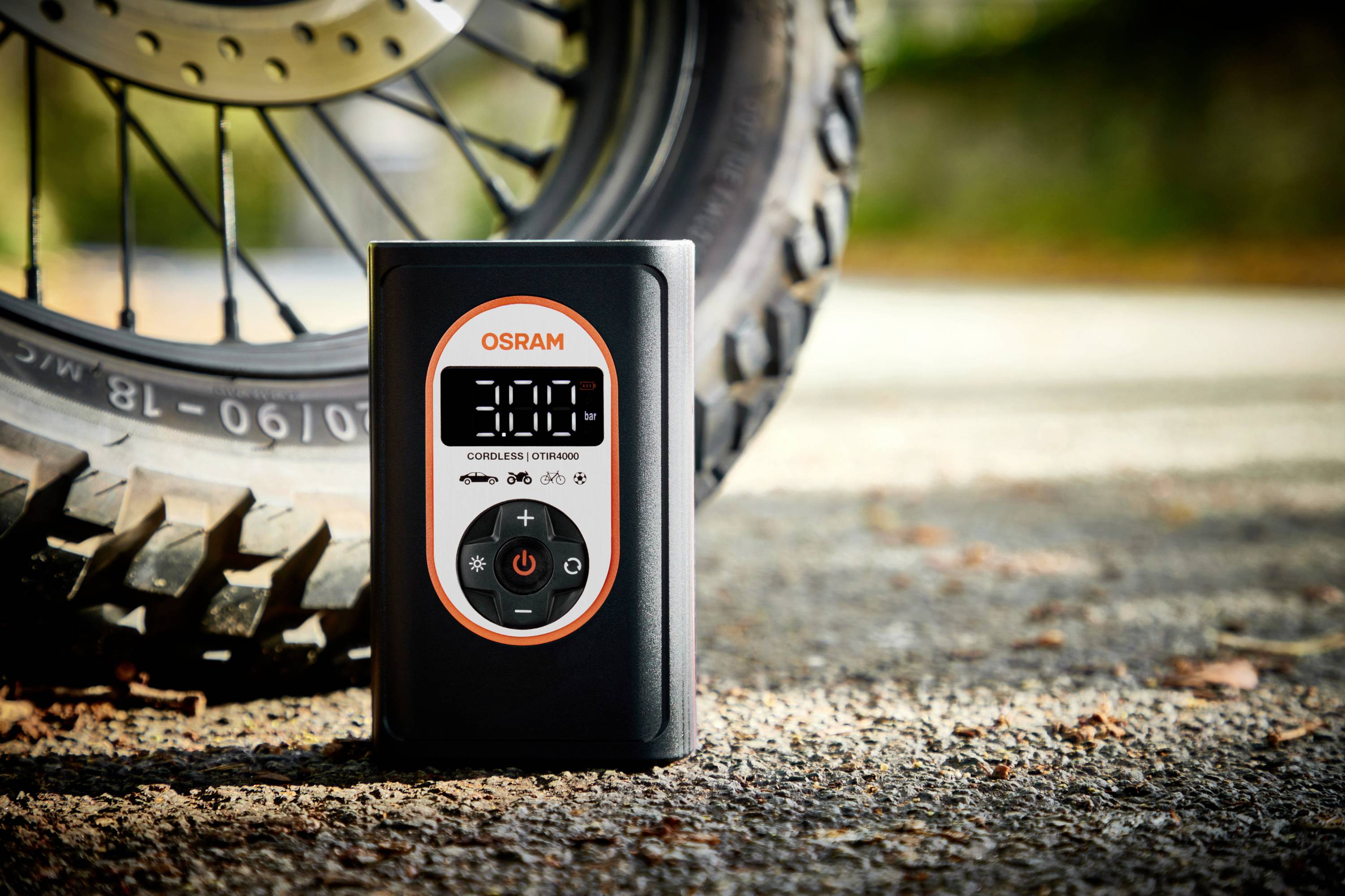 Cyfrowy miernik ciśnienia w oponach wskazuje '3.00' i stoi przed oponą motocykla na drodze.