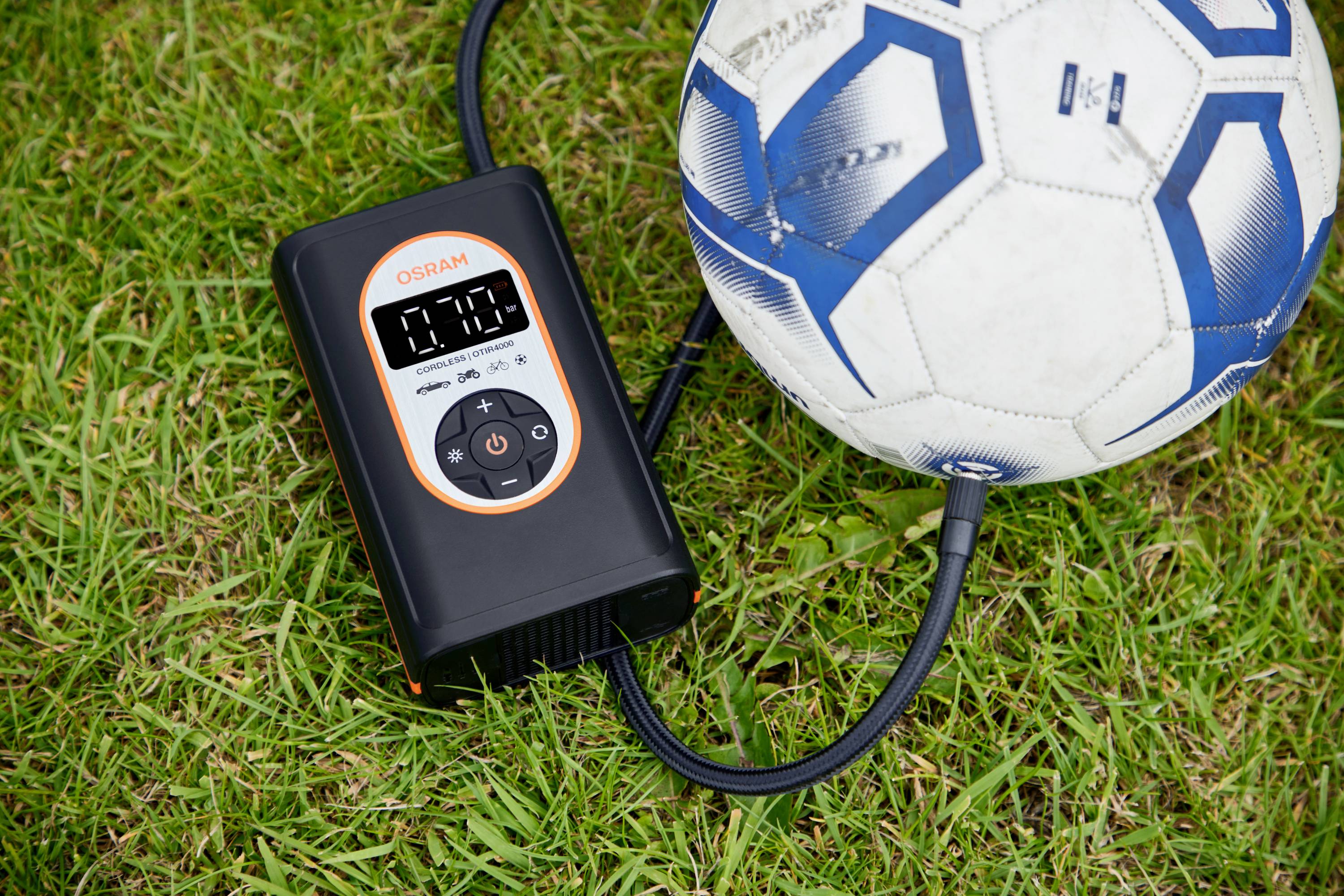Cyfrowy kompresor powietrza pompuje piłkę nożną. Na wyświetlaczu kompresora widać '0.28'. Oba przedmioty leżą na trawie.