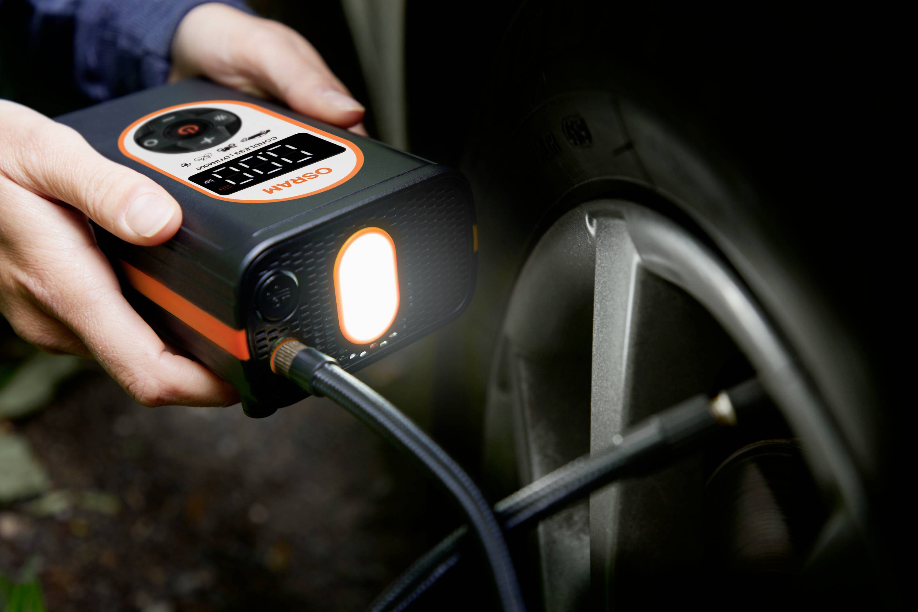 Osoba pompuje opony samochodu przenośnym kompresorem. Kompresor posiada wskaźnik ciśnienia.