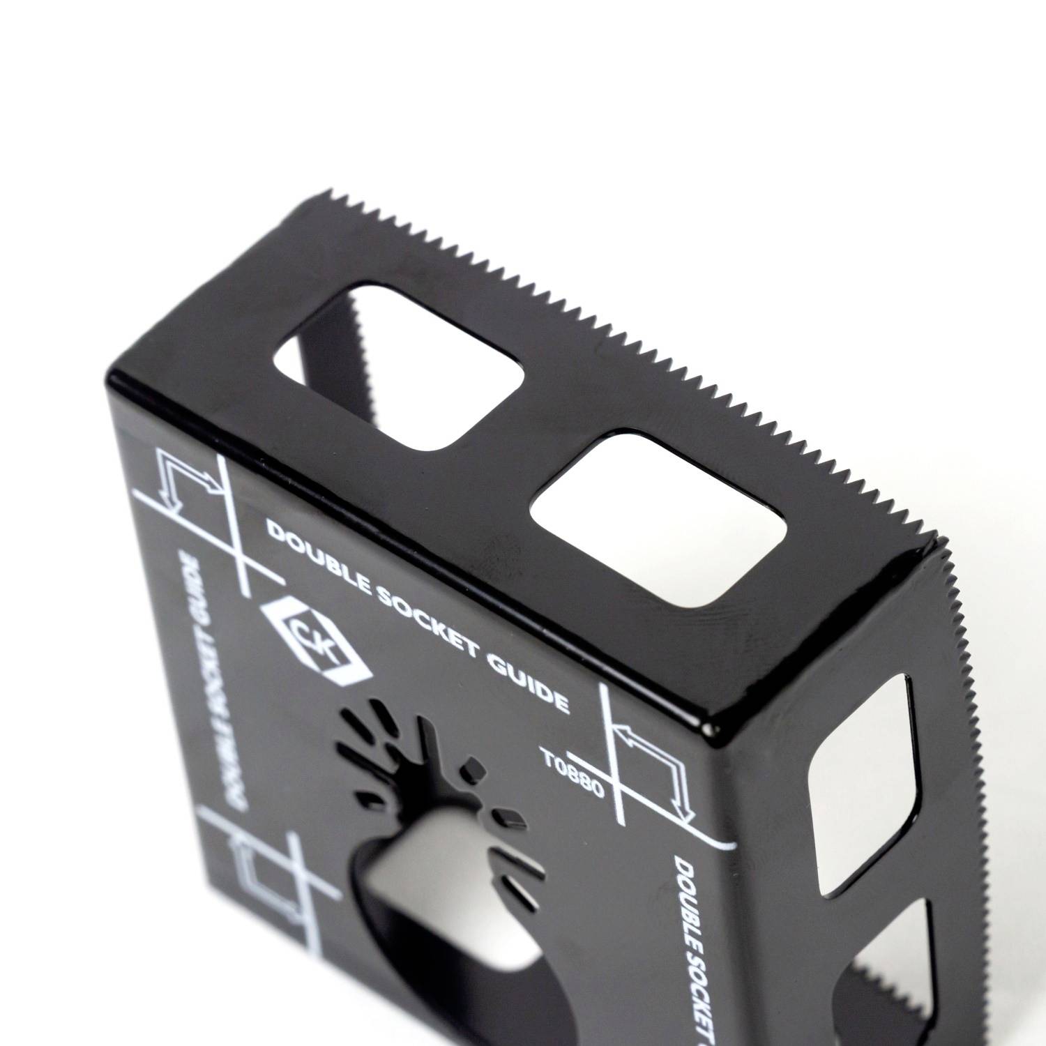 Czarne, kwadratowe narzędzie o ząbkowanych krawędziach i wyciętych prostokątach. Napis: 'Double Socket Guide'.