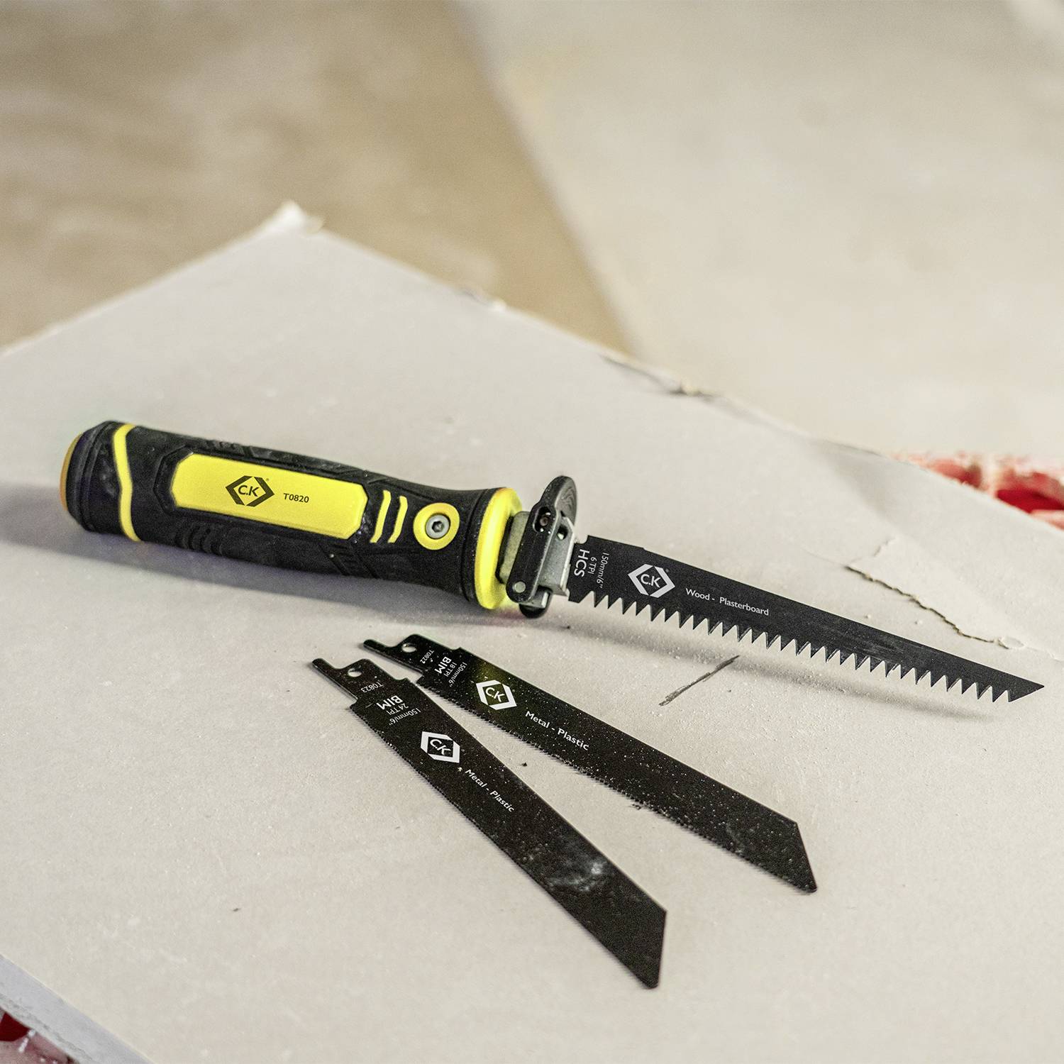 Żółta rękojeść z przymocowaną piłą tarczową leży na kawałku płyty gipsowo-kartonowej. Obok niej znajdują się trzy dodatkowe ostrza piły.