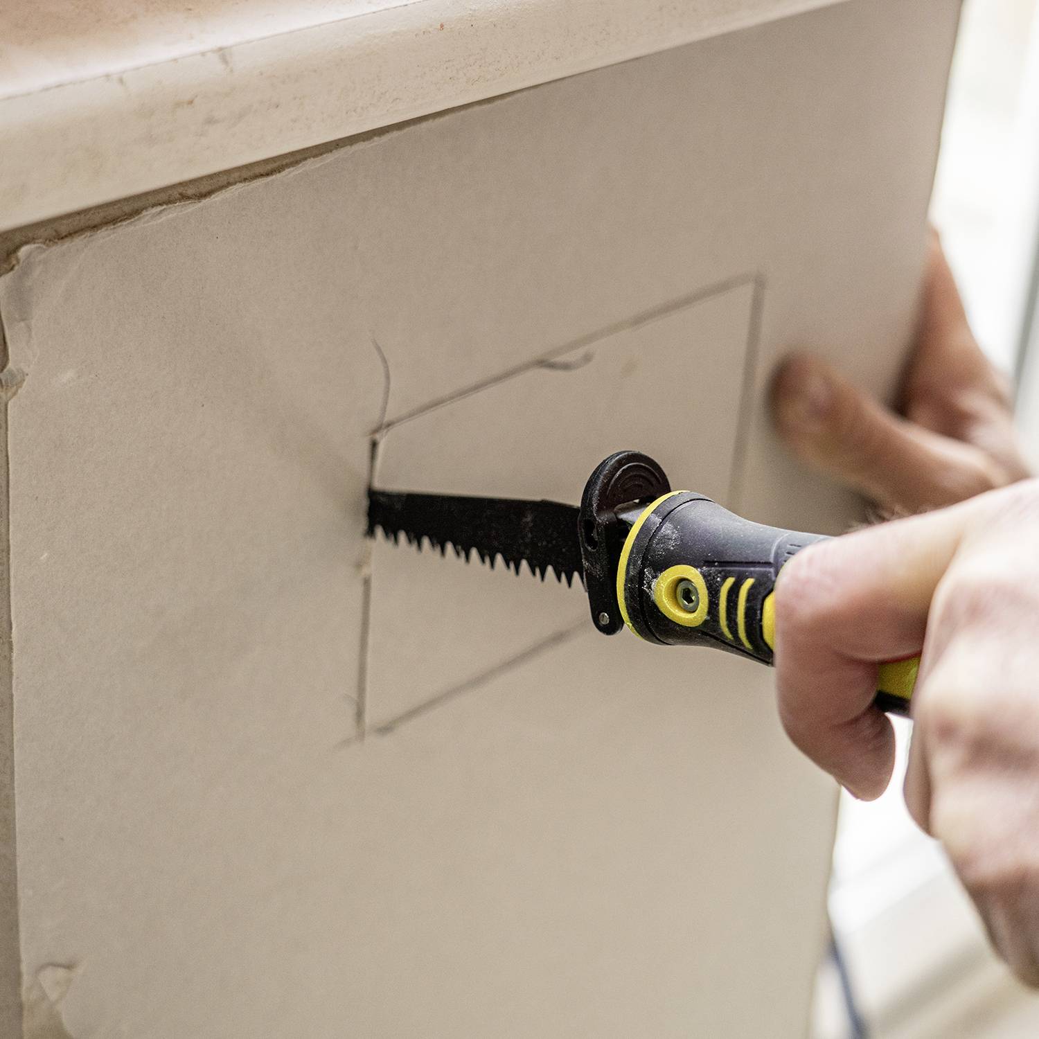 Osoba wycina piłą kwadratowy otwór w ścianie z płyty gipsowo-kartonowej. Zbliżenie na ręce, skoncentrowana czynność.